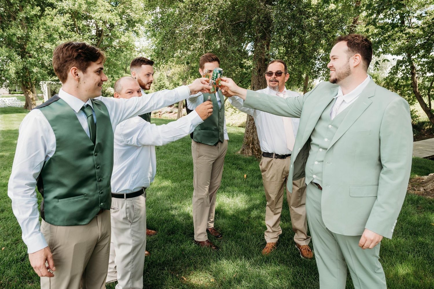 Groom and groomsmen in sage green suits toasting outdoors at a wedding celebration.