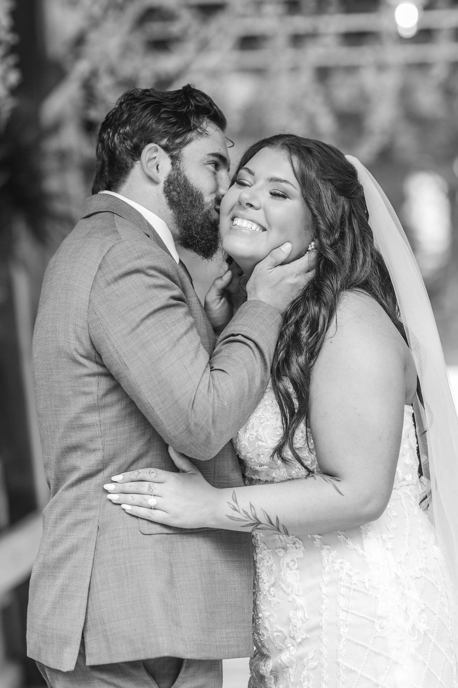 Black and white photo of a groom kissing his bride on the cheek as she smiles joyfully on their wedding day.
