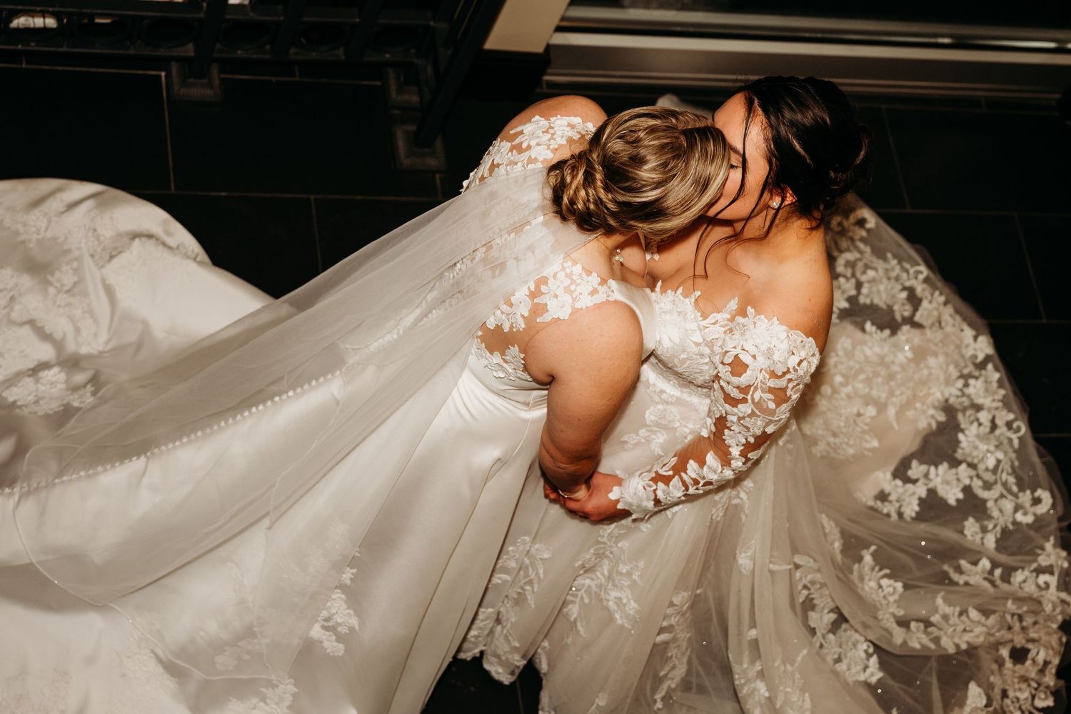 A bride in an elegant lace wedding dress with flowing veil walks down a dimly lit hallway.