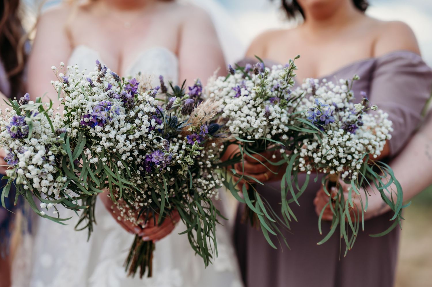 Wedding bouquets featuring delicate white baby's breath and purple wildflowers with eucalyptus leaves held against mauve dresses.