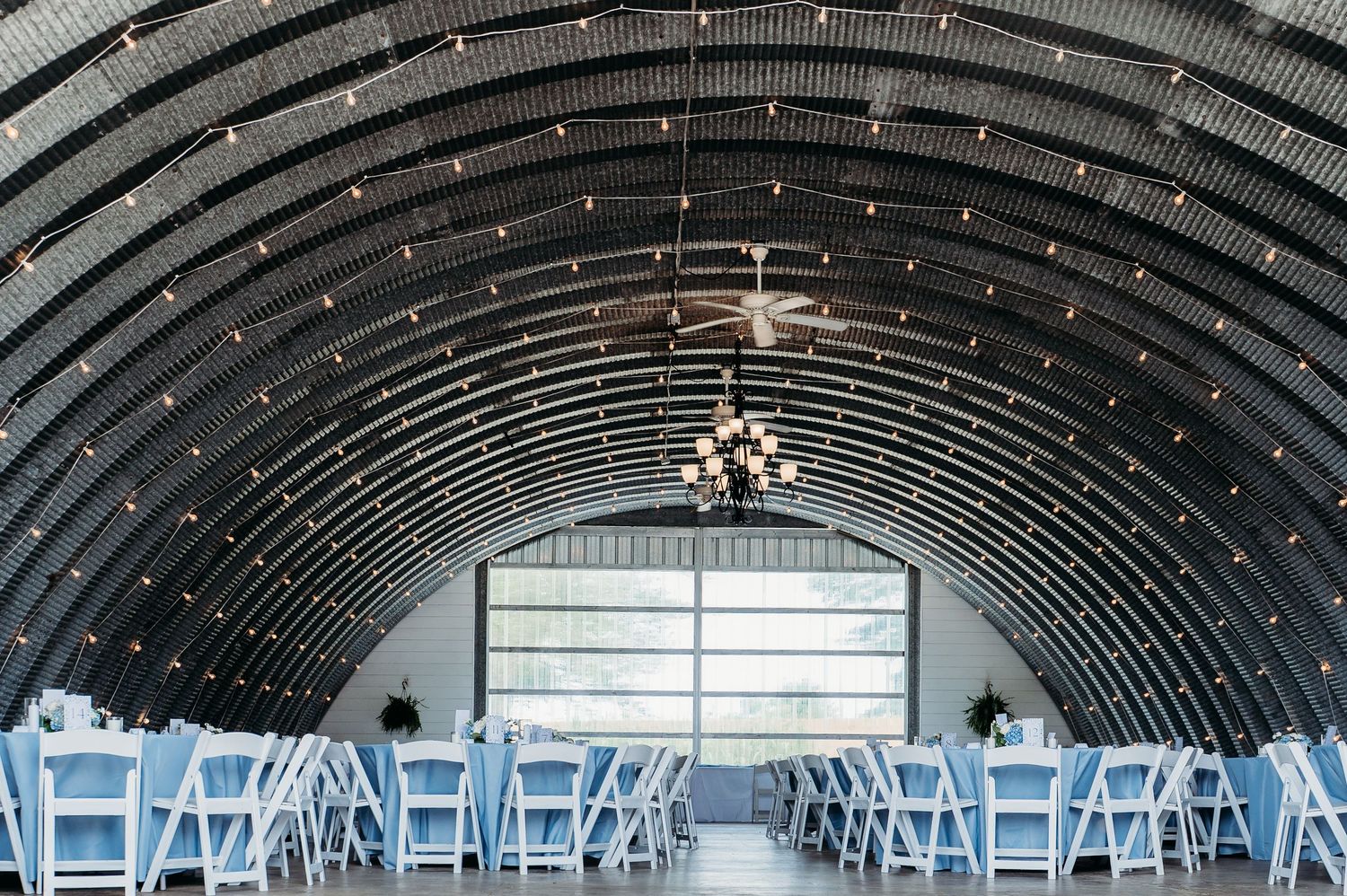 Elegant wedding venue inside an arched barn with string lights, chandelier, and white chairs arranged for a ceremony.