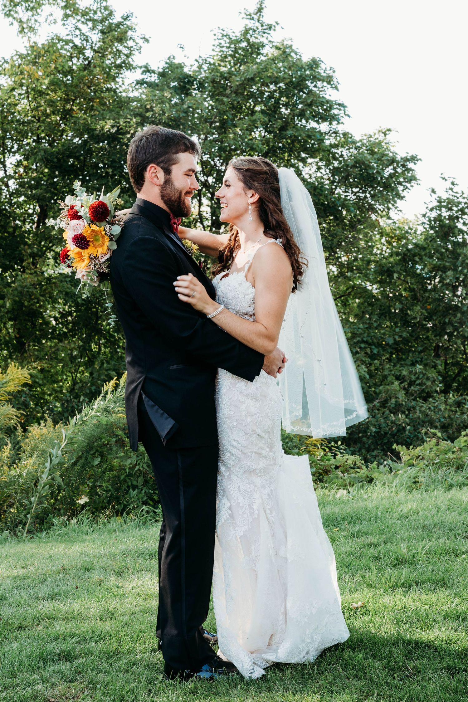 Bride and groom embrace outdoors, smiling at each other, she in lace gown with veil, he in black tuxedo, holding bouquet.