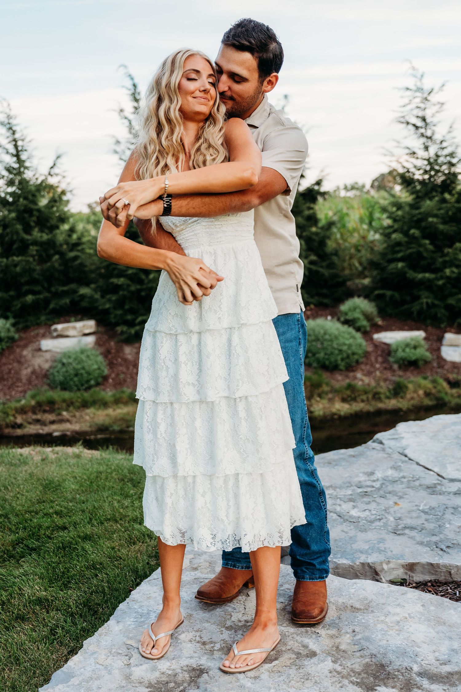 A couple embraces on a rock outdoors, with the person wearing a white tiered lace dress and sandals against a garden backdrop.
