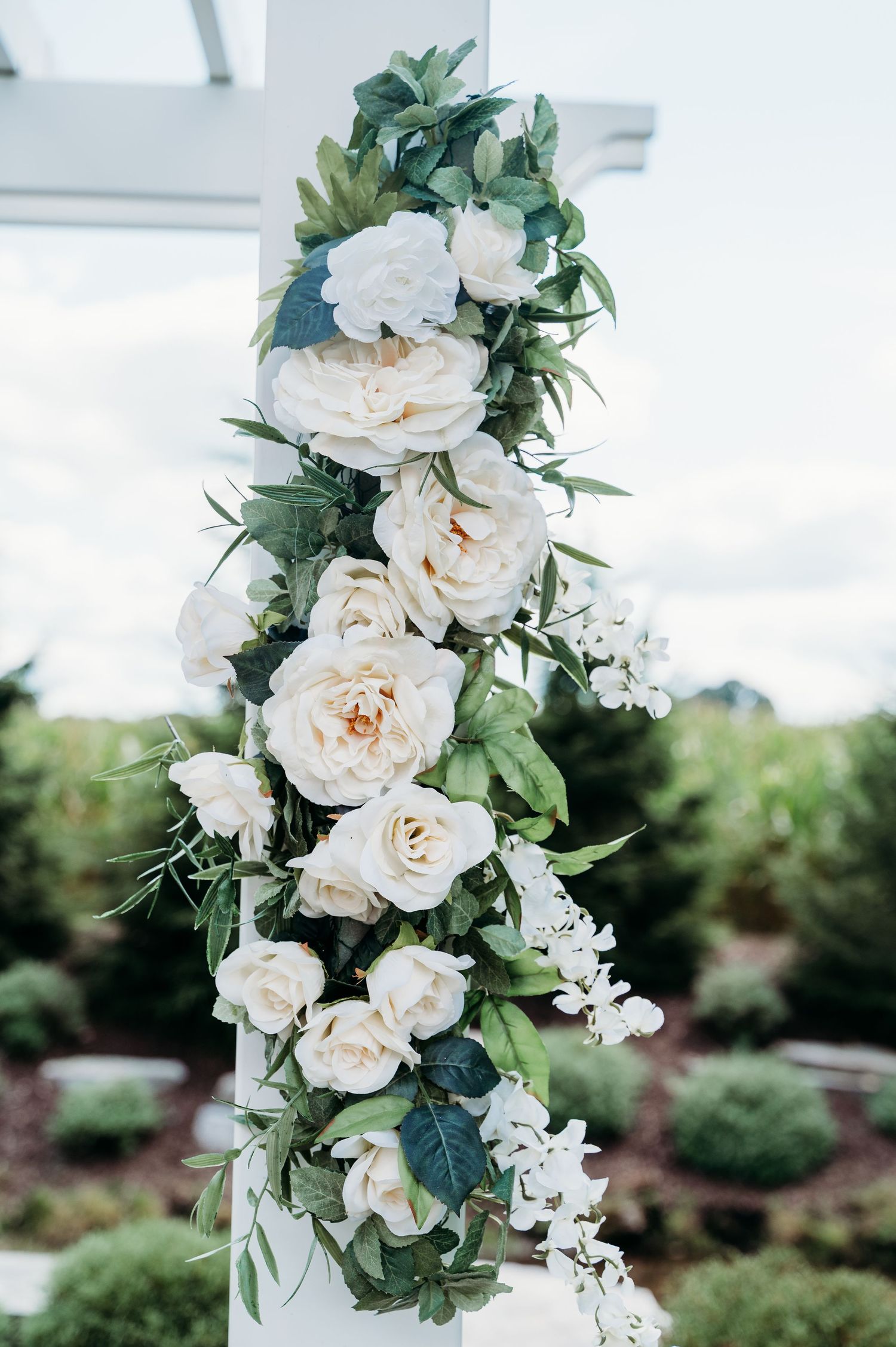 Elegant white roses and greenery arranged in a cascading floral wedding arch decoration against an outdoor garden backdrop.