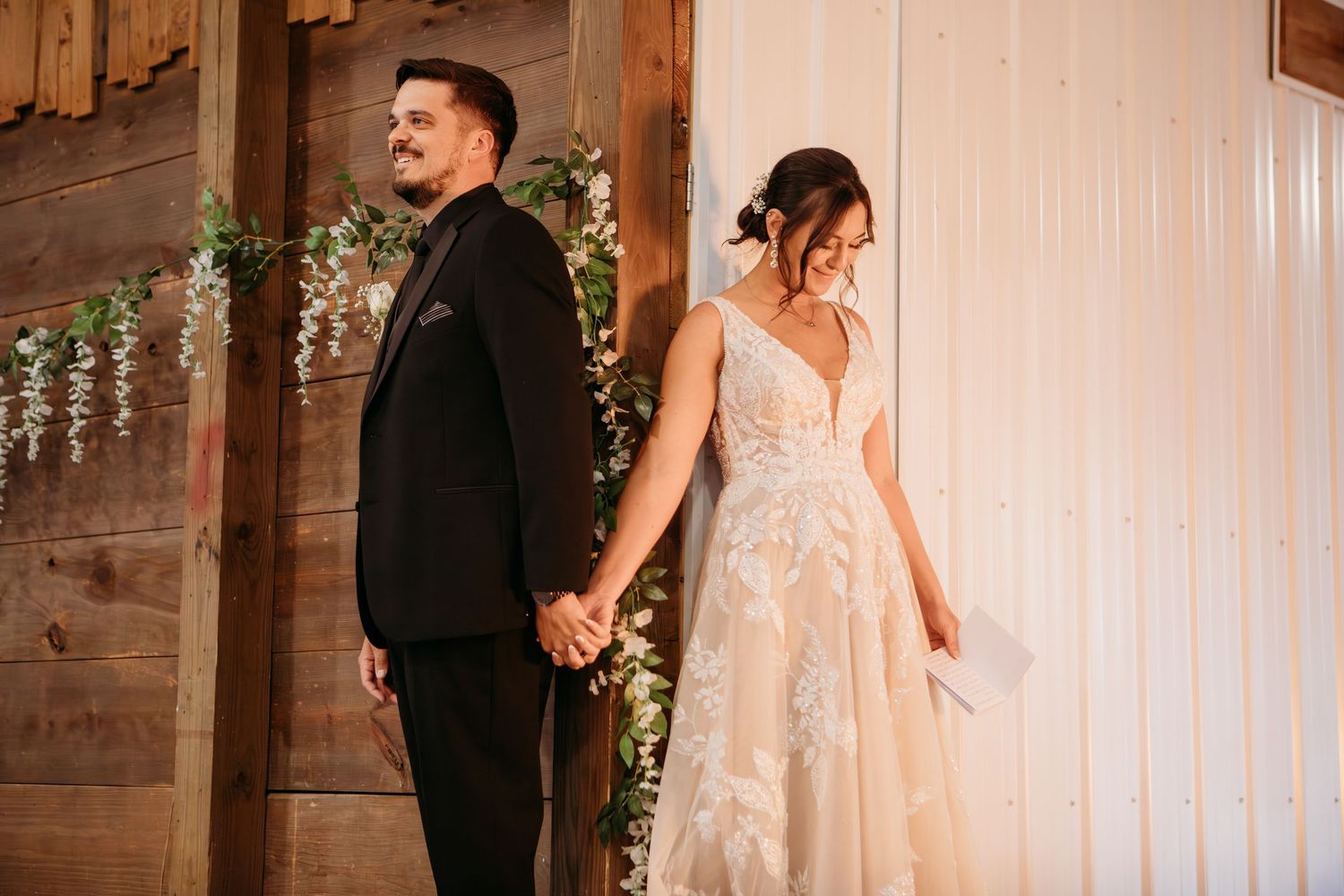 A newlywed couple holds hands at their rustic barn-style wedding venue decorated with delicate floral garlands.
