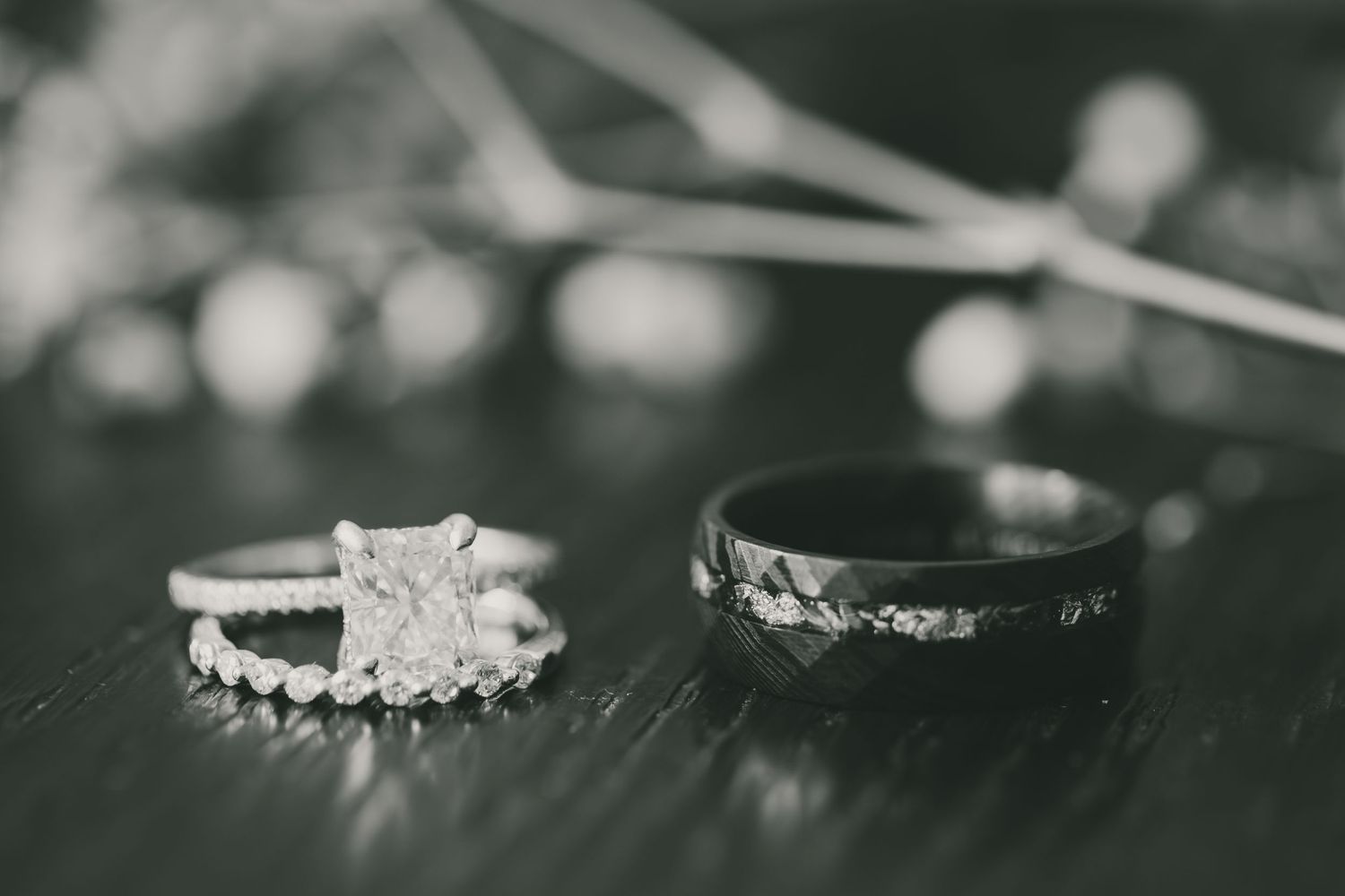 Black and white close-up of diamond engagement ring and wedding band set with sparkly bokeh in background.