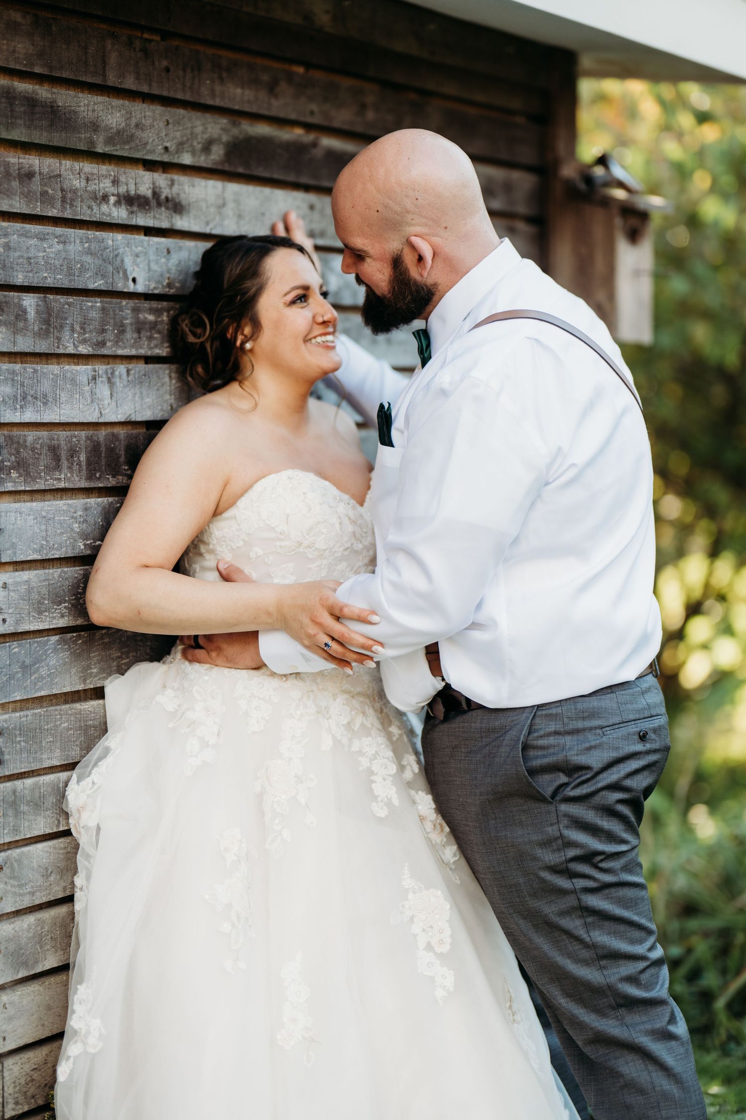 Newlywed couple share a tender moment while standing against rustic wooden wall backdrop during wedding photoshoot.