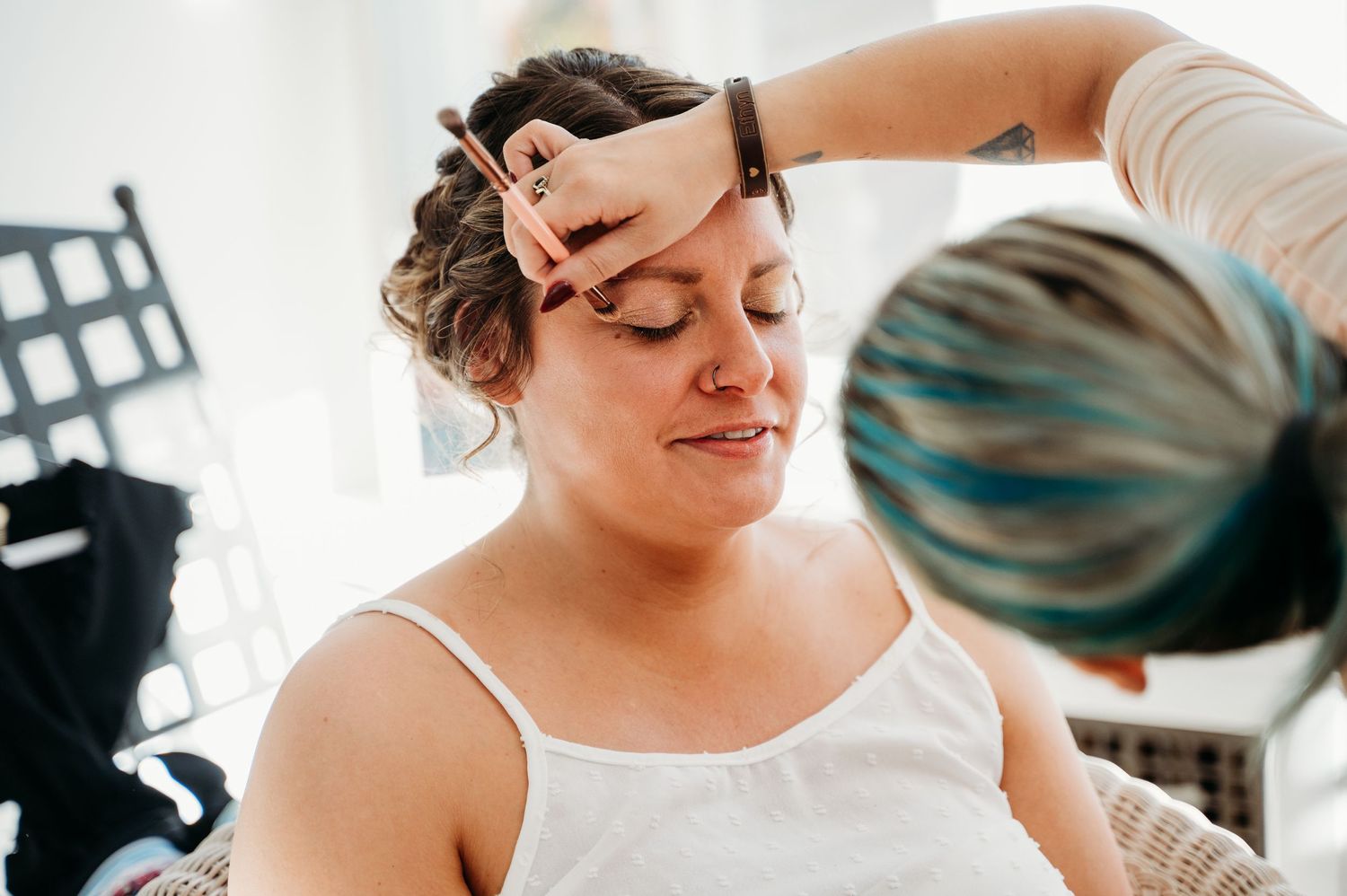 Makeup artist carefully applies cosmetics to client wearing white camisole during professional beauty session.