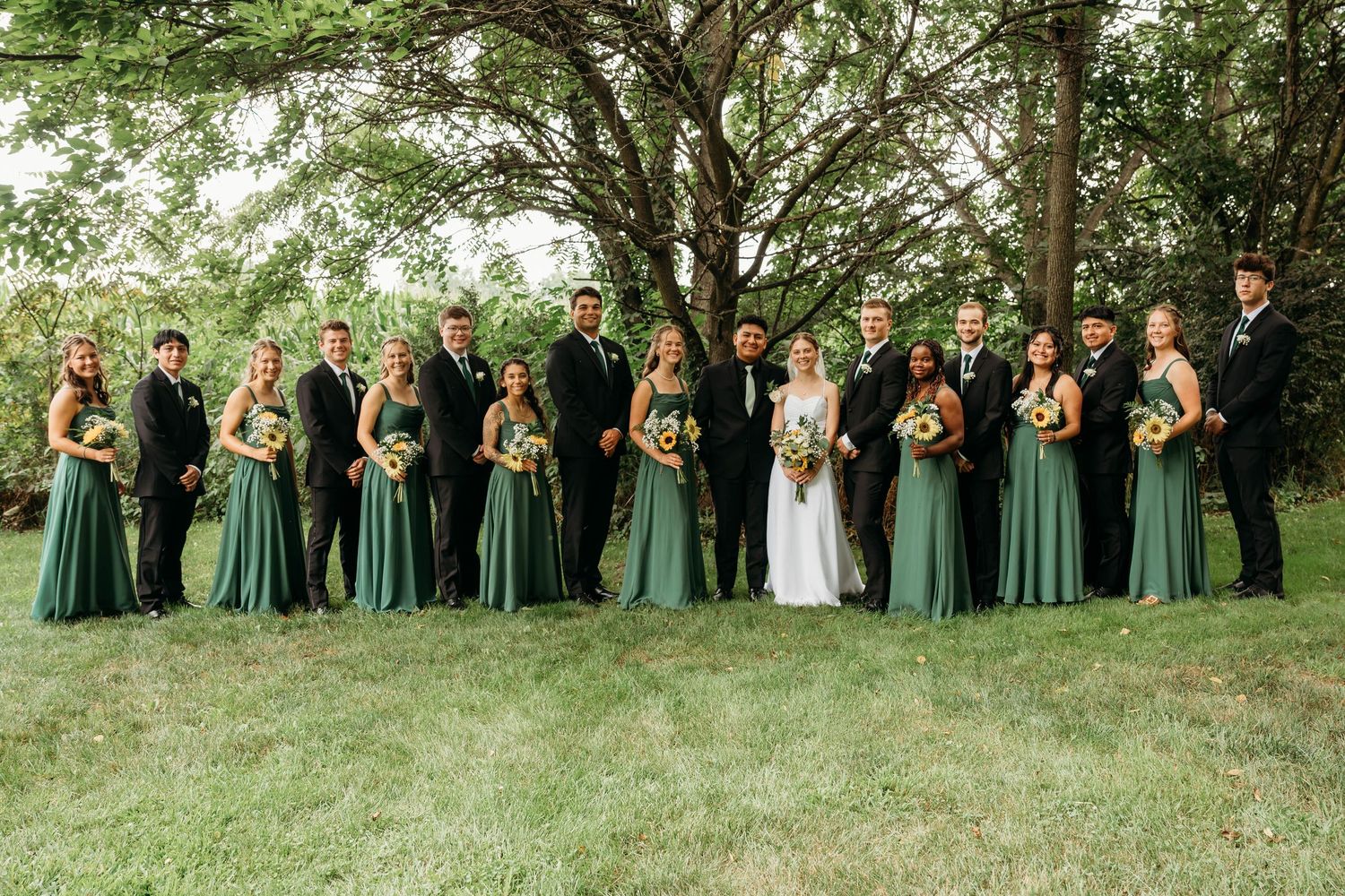 Wedding party in sage green dresses and black suits posing outdoors under trees on a lush green lawn.