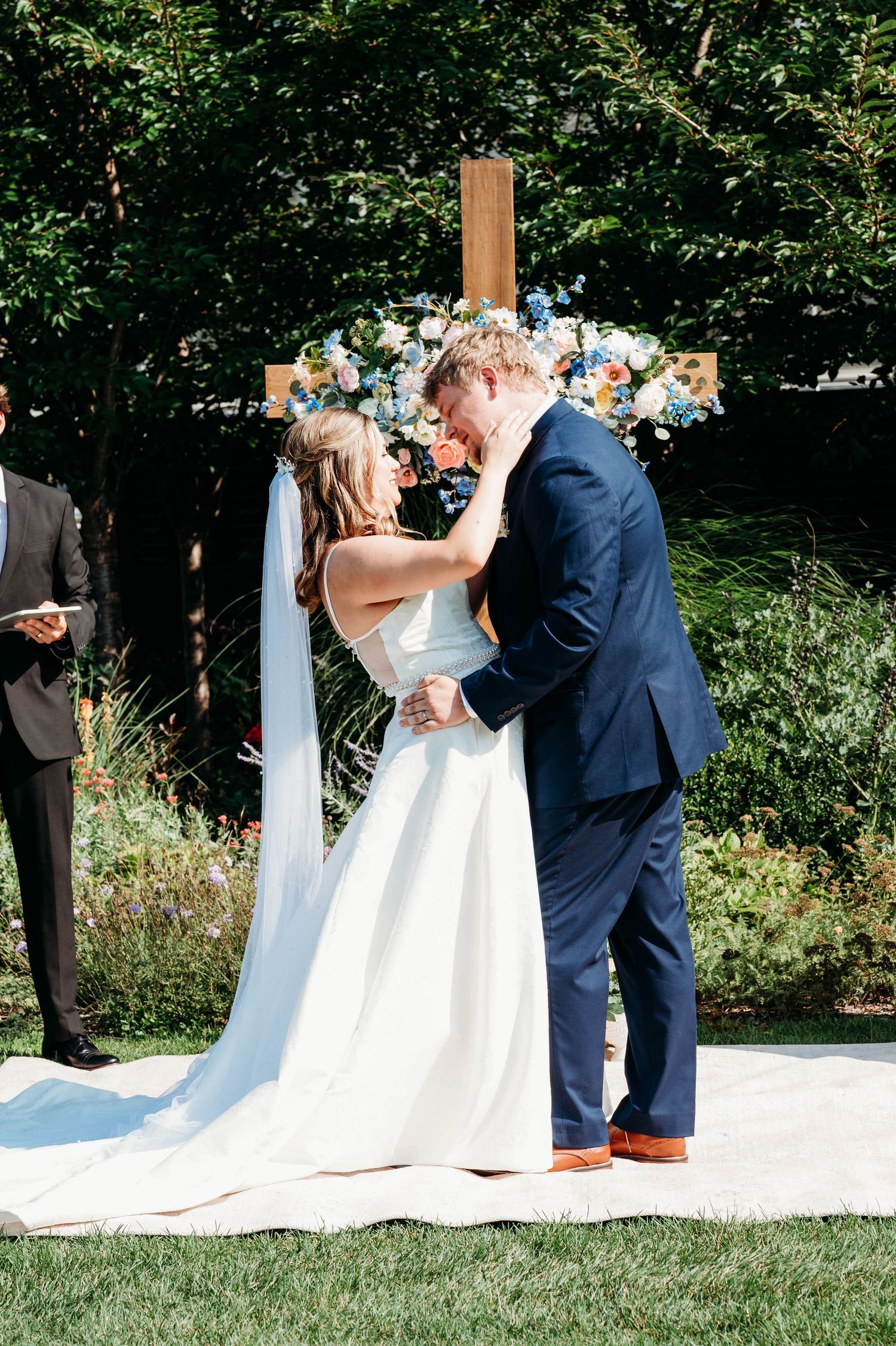 Bride and groom share first kiss at outdoor wedding ceremony before floral cross altar.