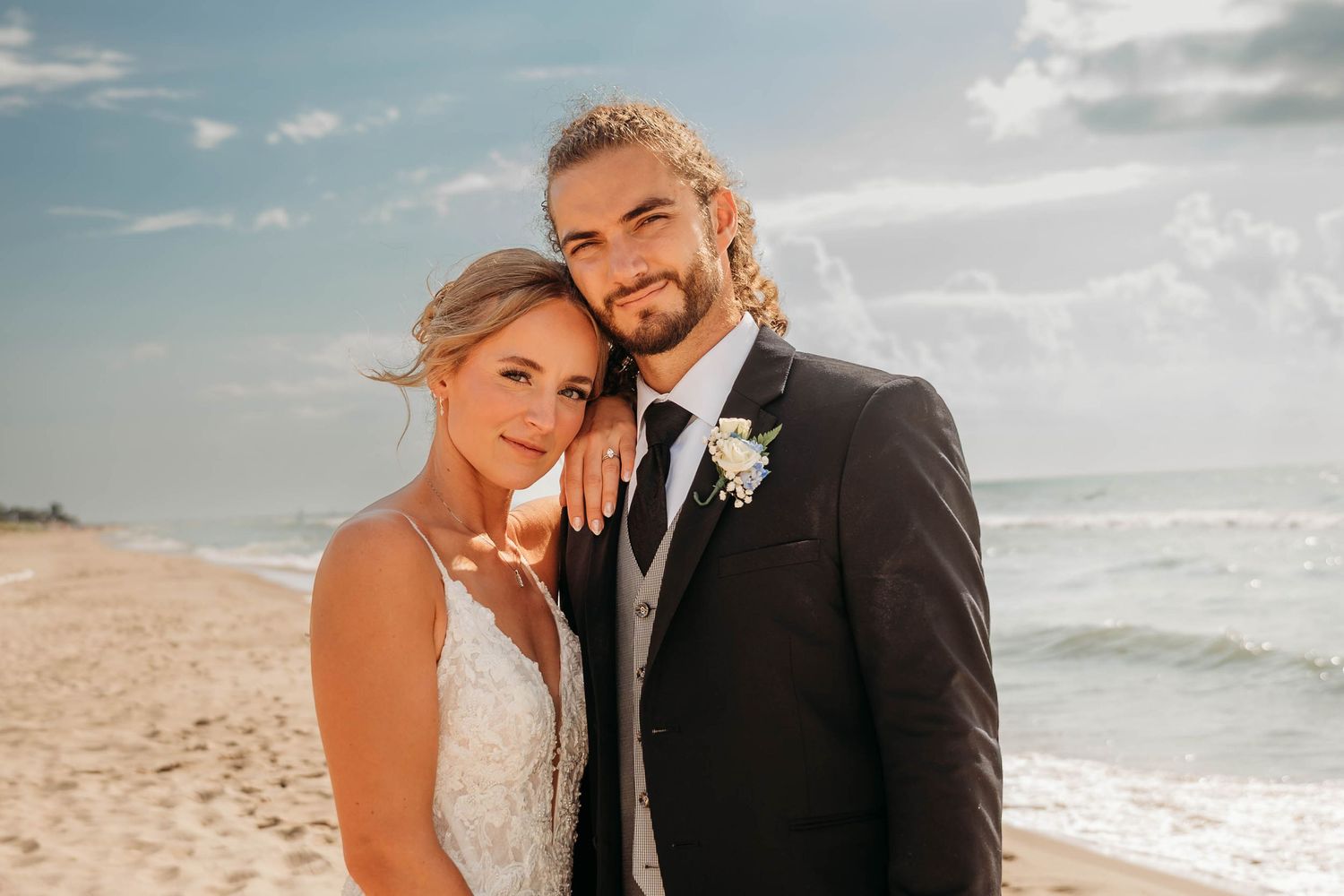 Bride and groom smiling together on a sandy beach, groom in dark suit with boutonniere, bride in white lace dress.