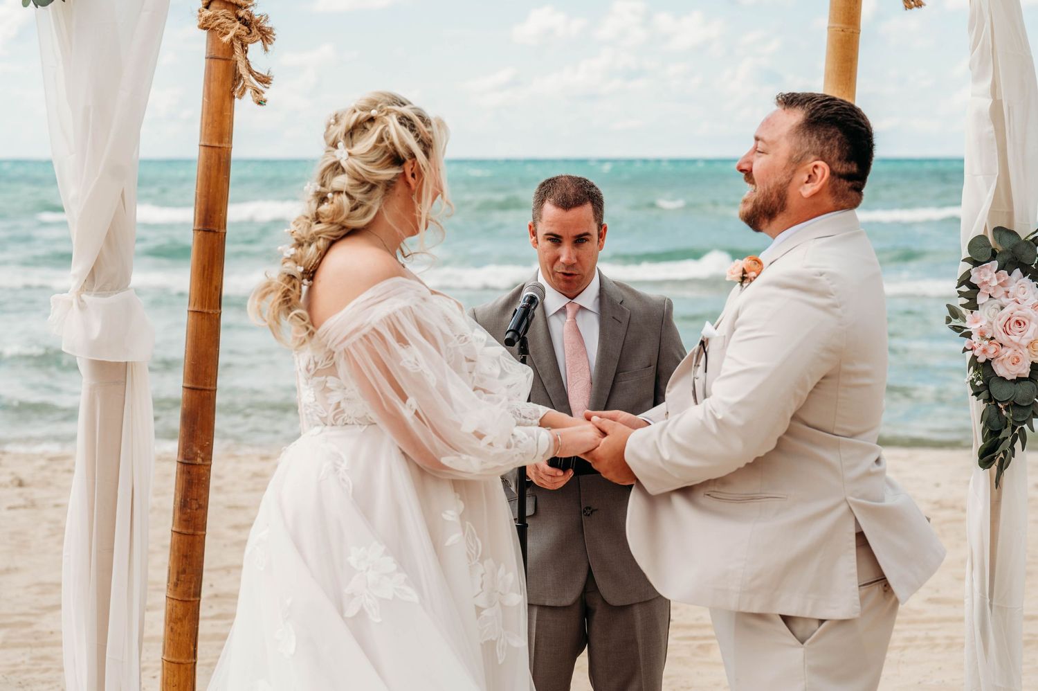 A wedding celebration on a beach with white fabric draping and floral arrangements under bright sunny skies.