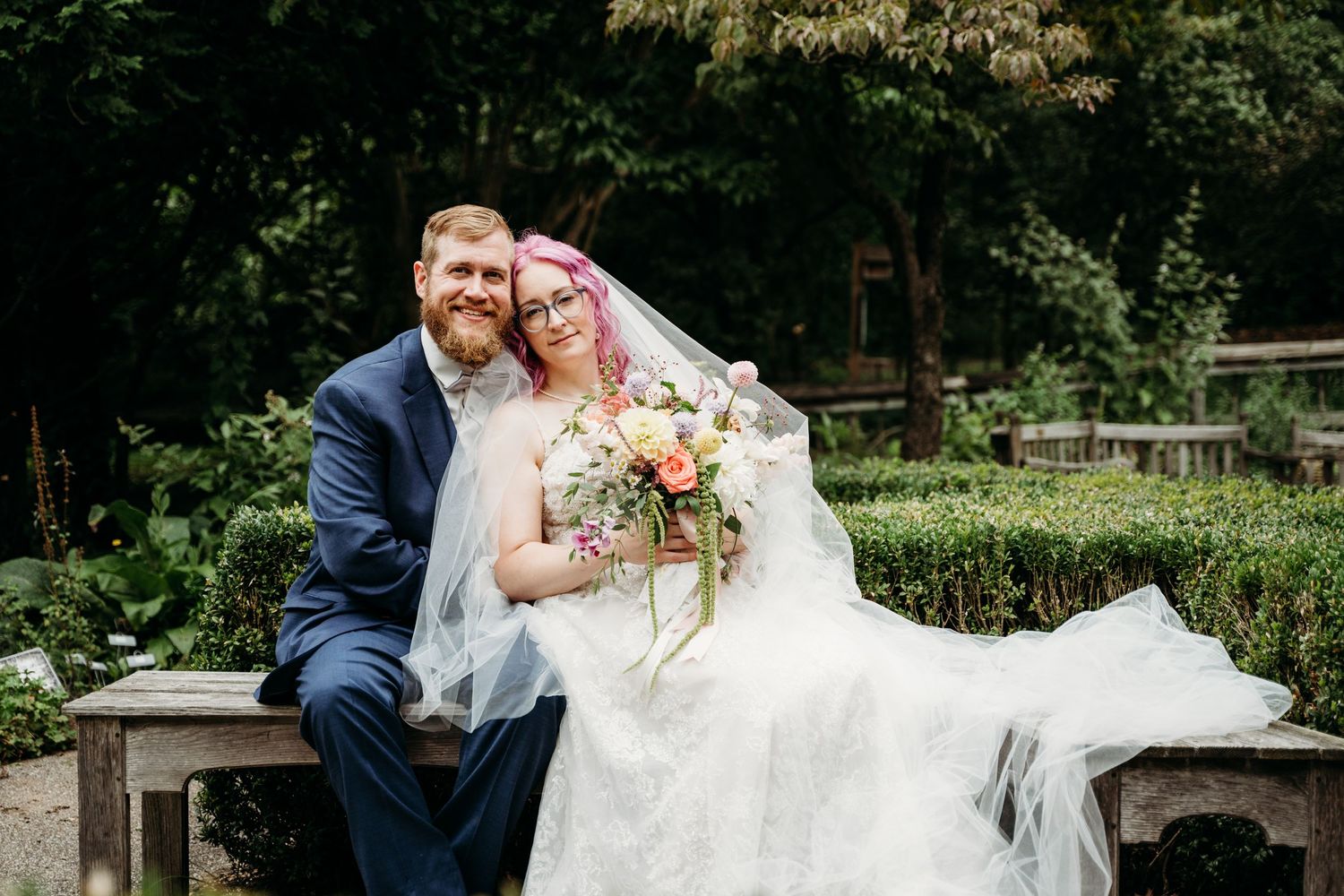 A romantic wedding couple sits on stone steps in a garden setting, surrounded by greenery and holding a floral bouquet.