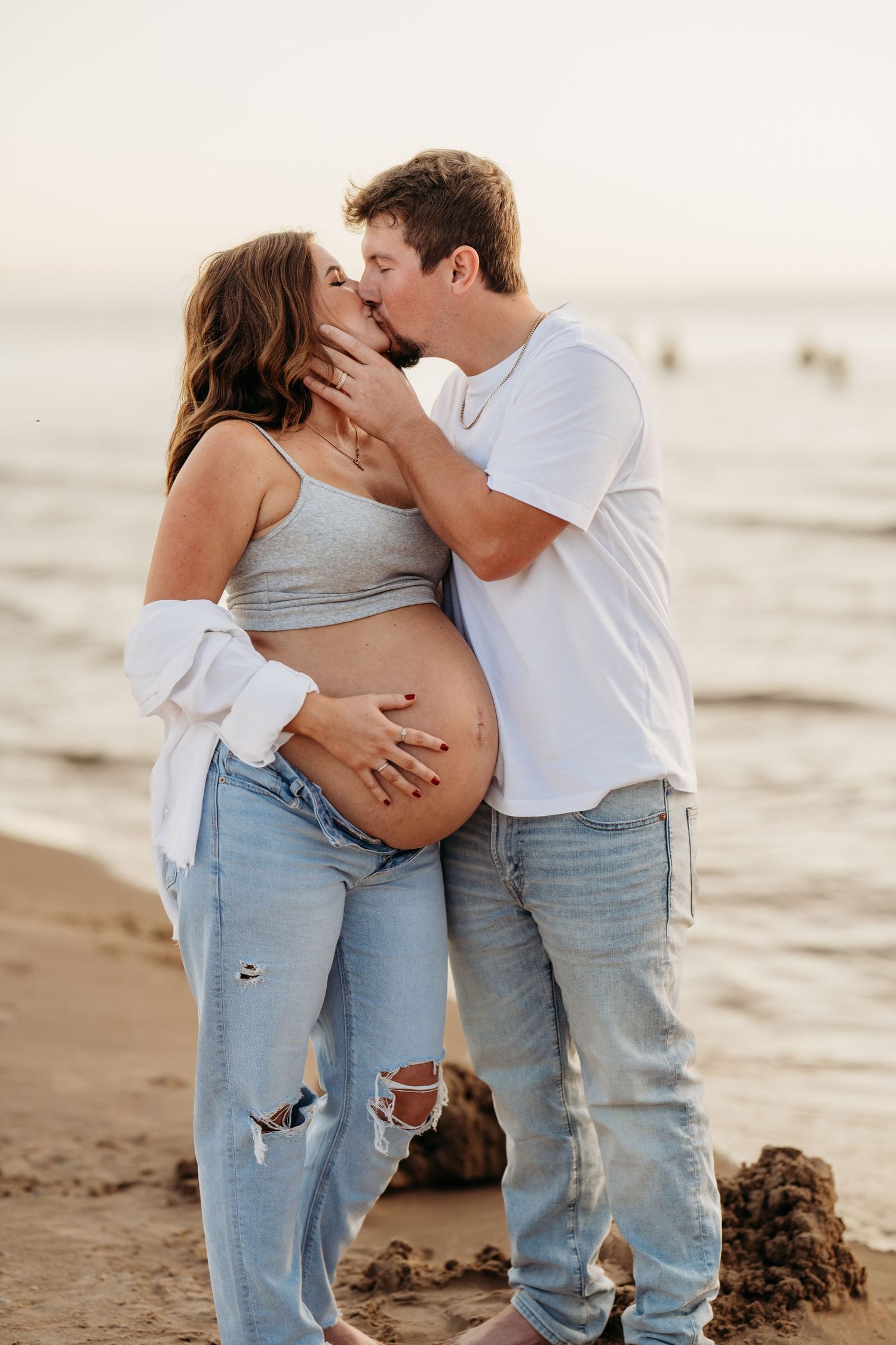 A couple shares an intimate moment at sunset on the beach while cradling a pregnant belly in casual attire.