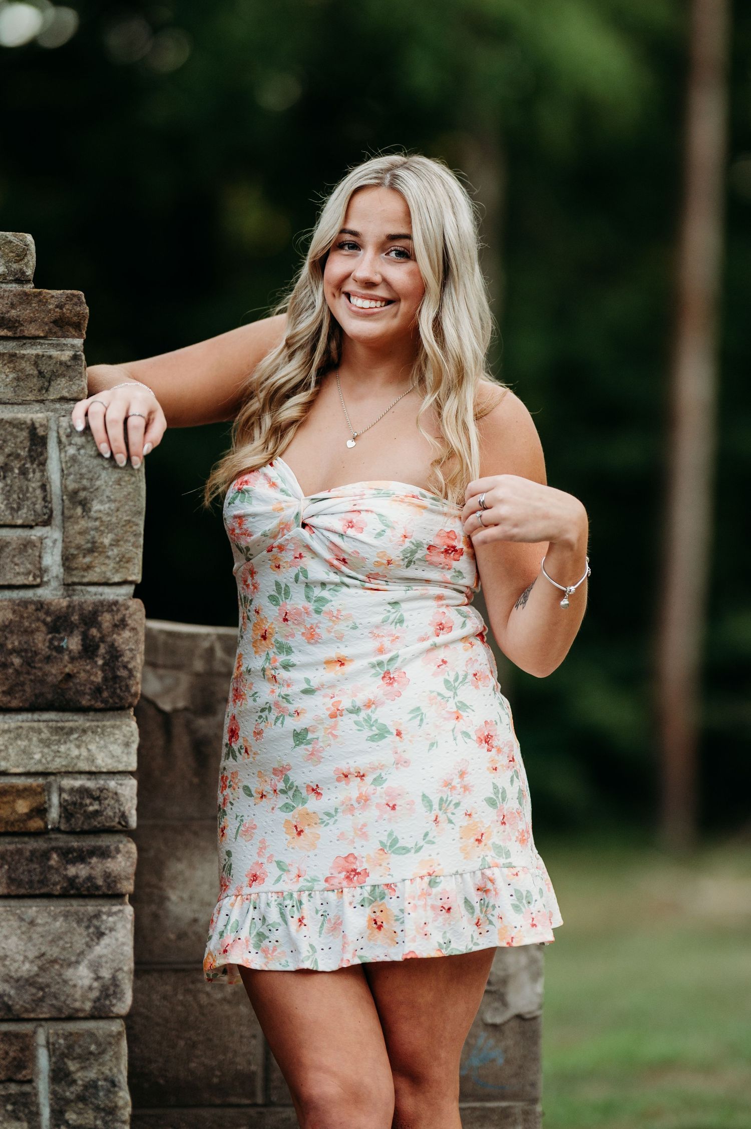 A floral print summer dress is showcased outdoors against a stone wall backdrop in natural lighting.
