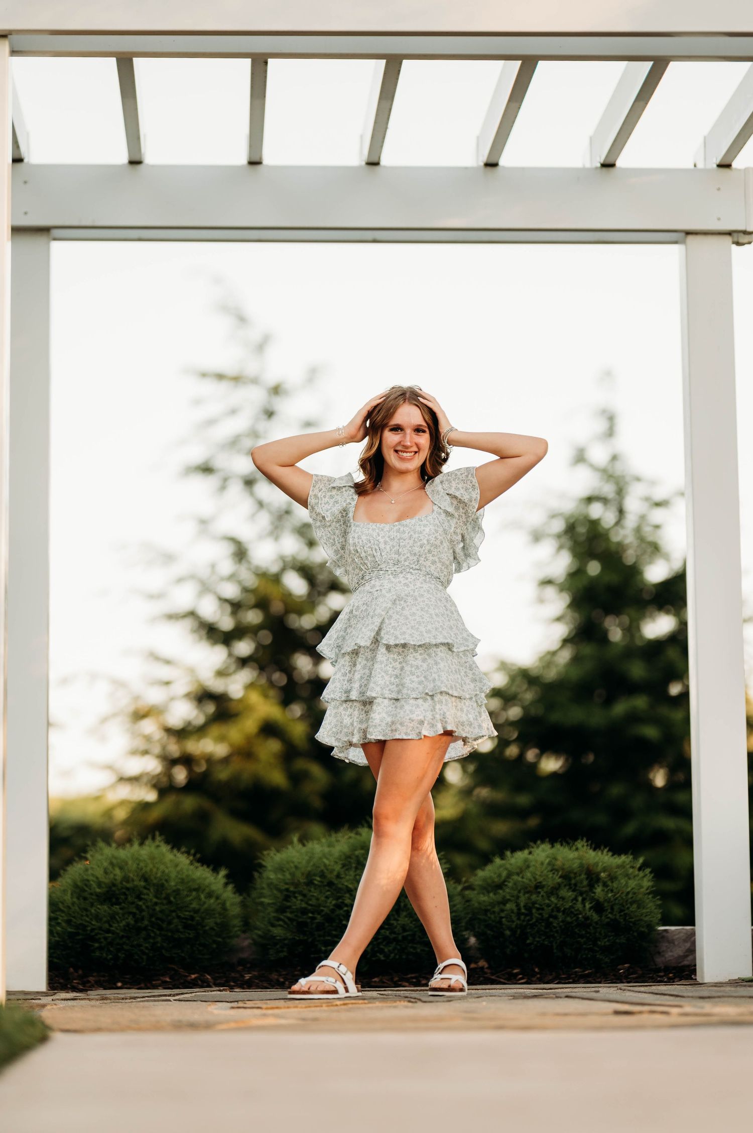 Young woman in a floral ruffle mini dress poses under a white pergola surrounded by lush greenery.
