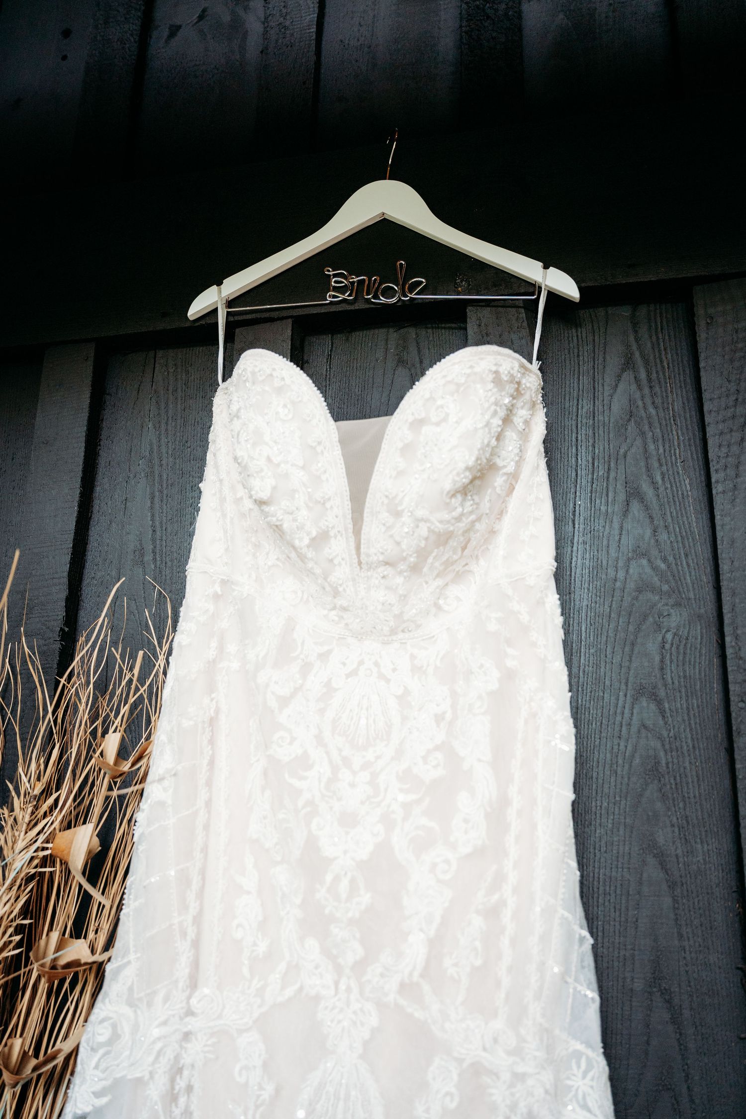 White lace strapless wedding gown hanging on a Bride hanger against a dark wood wall.