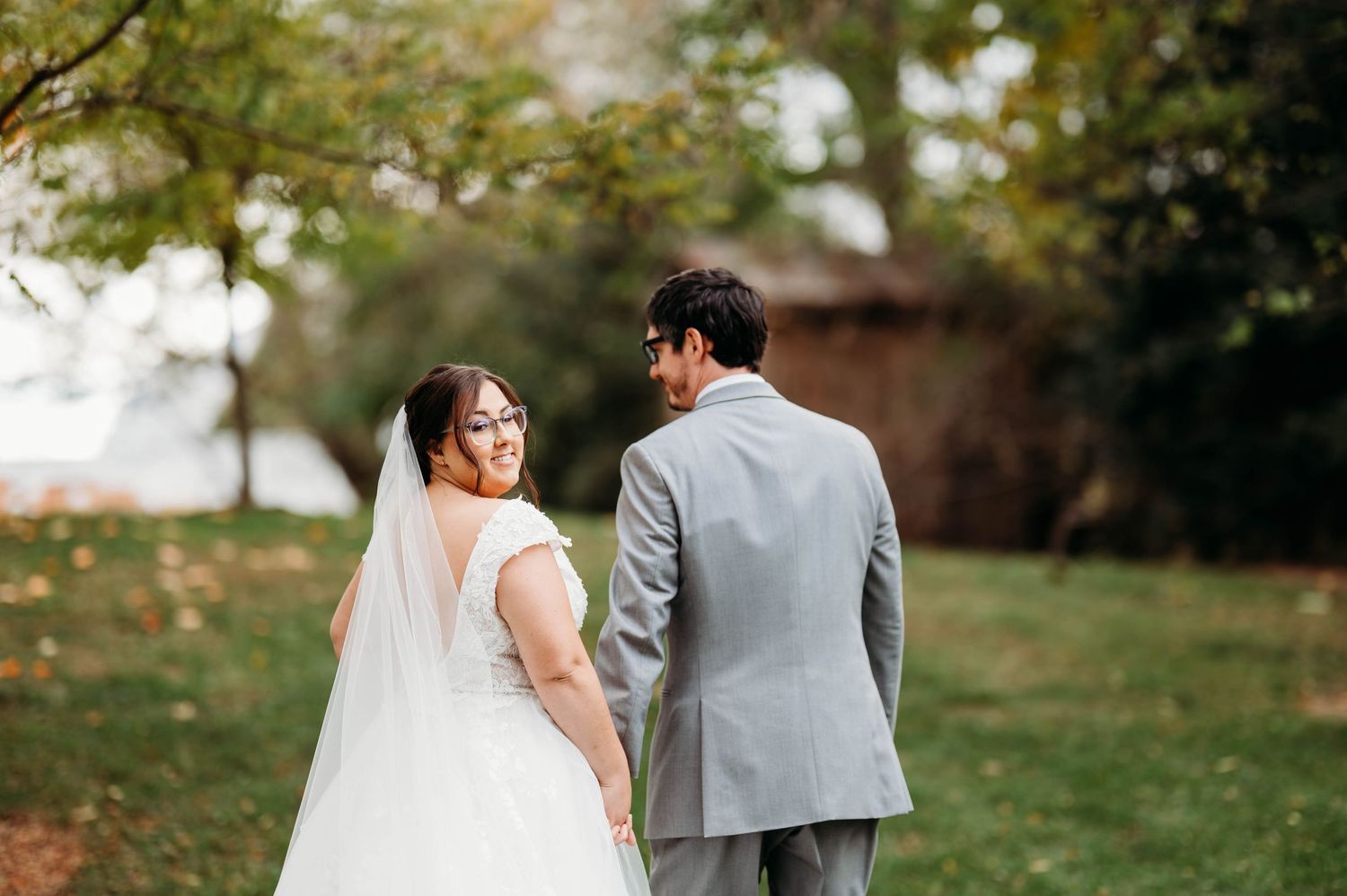 Bride in white gown and veil walks with groom in gray suit outdoors on lush green lawn at wedding.