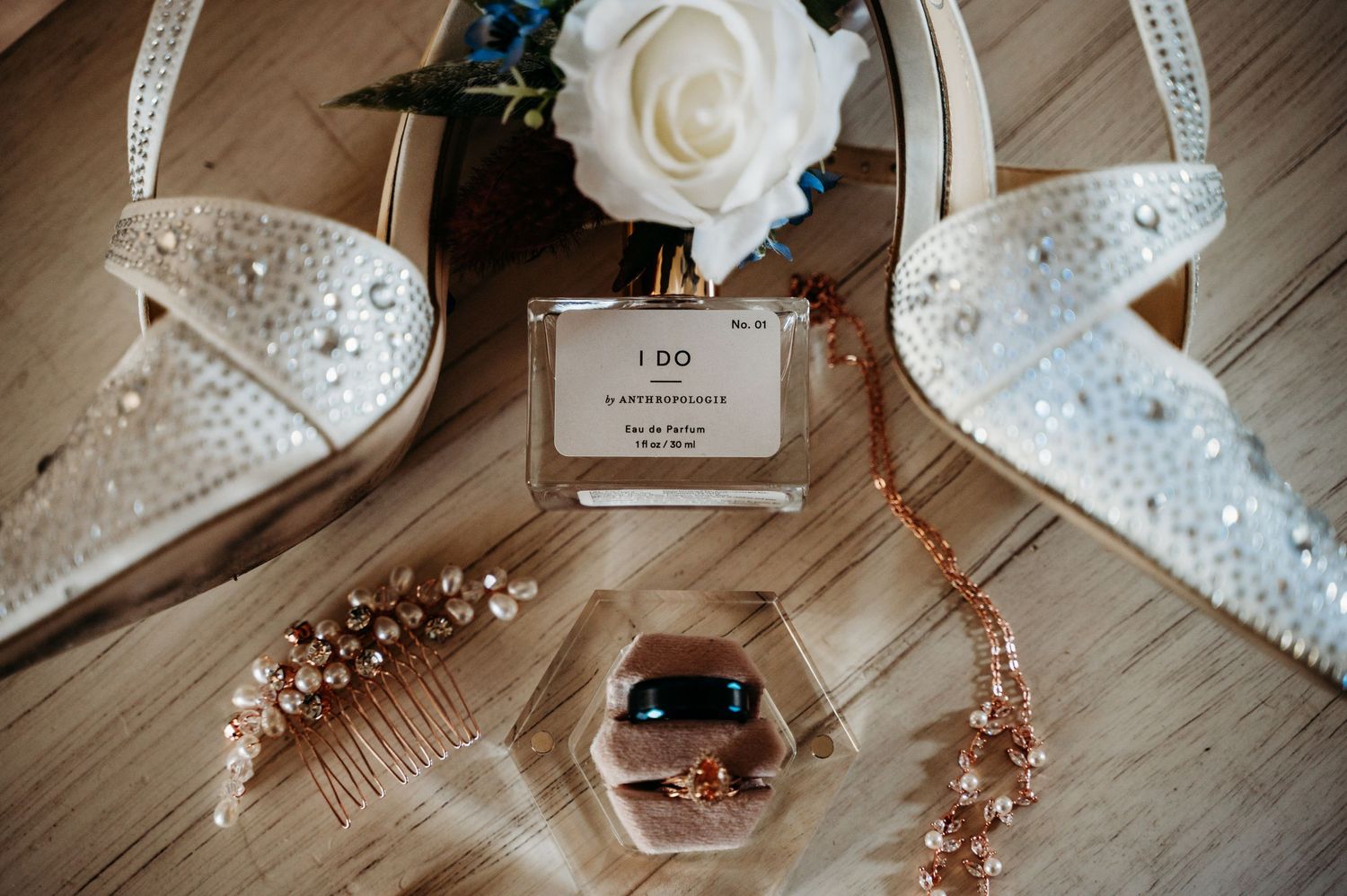 Wedding accessories including sparkly white shoes, perfume bottle, pearl jewelry and a white rose on wooden surface.