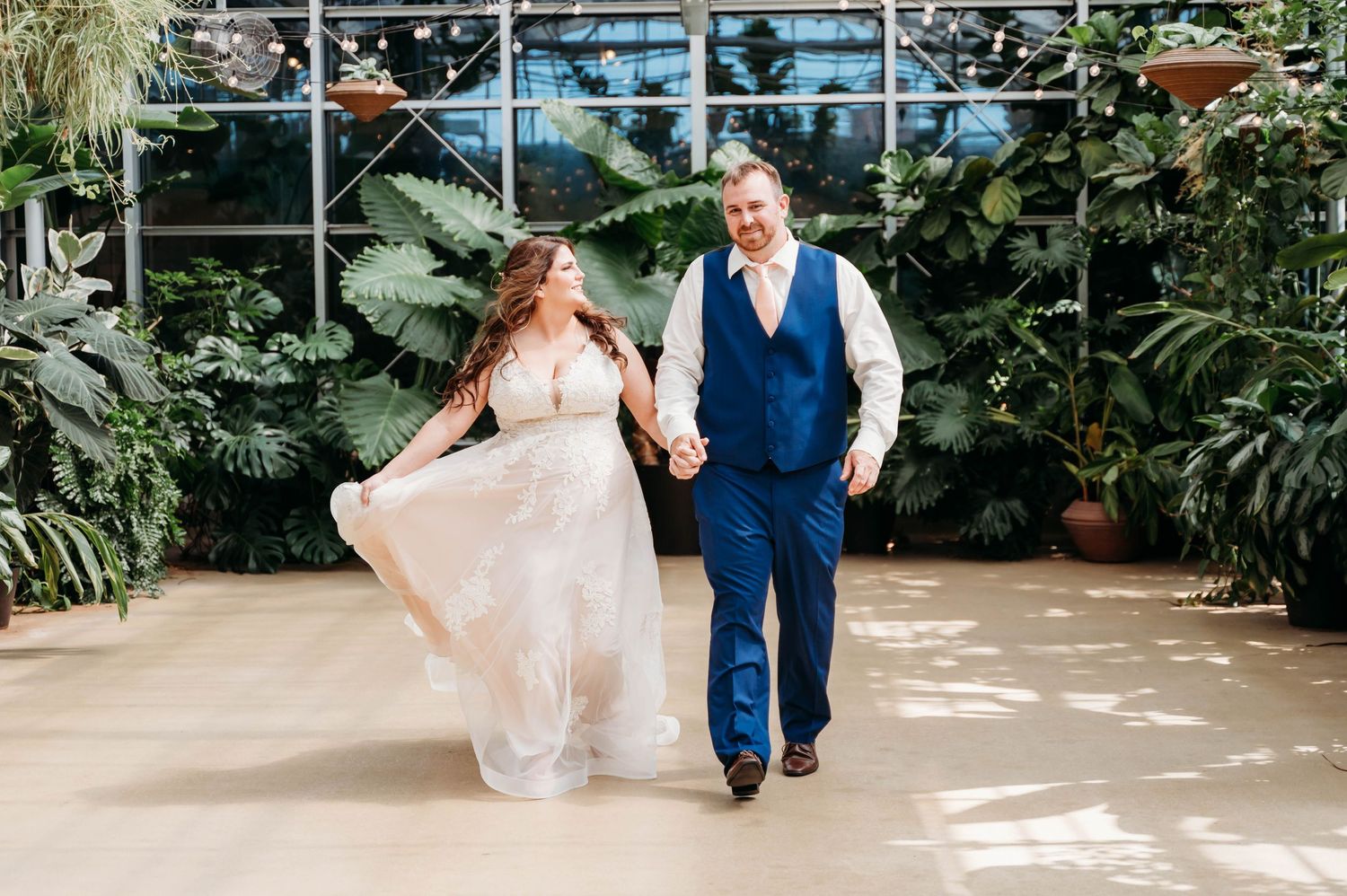 A couple walks hand in hand through a greenhouse conservatory, with the bride wearing a flowing blush gown.