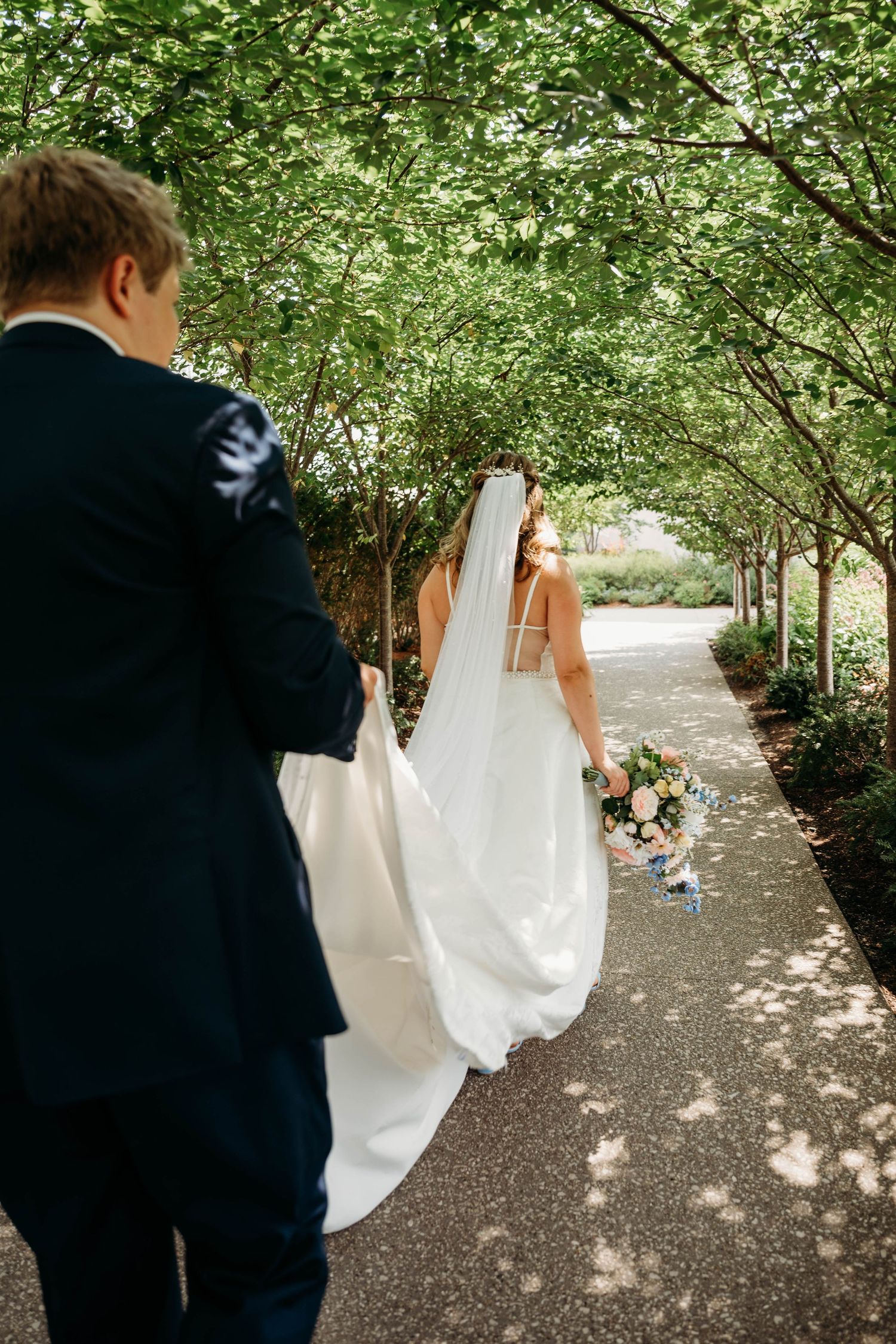 Bride and groom walking through a tree-lined garden path, bride holding a floral bouquet in a white gown with veil.
