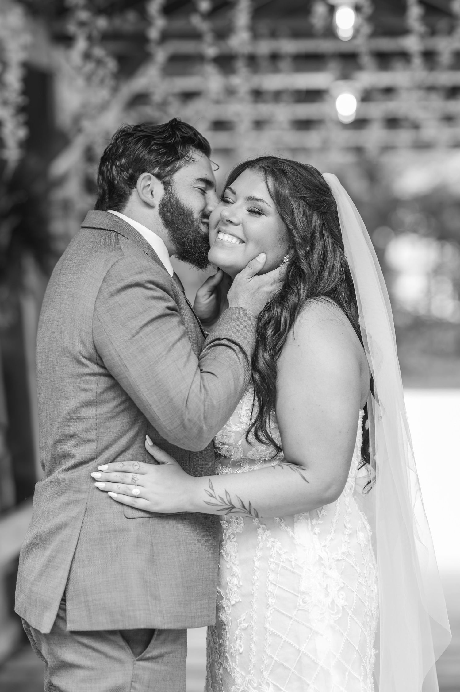 Black and white wedding photo of groom kissing bride on cheek as she smiles, wearing veil and lace gown.
