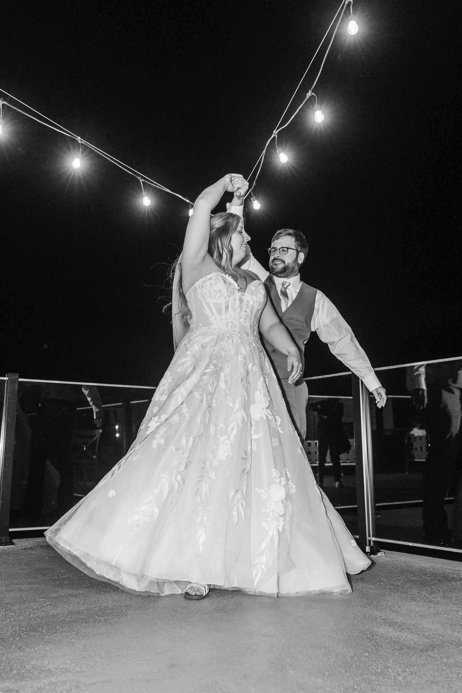 A couple dances together at their wedding reception, with the bride's white dress twirling under string lights at night.