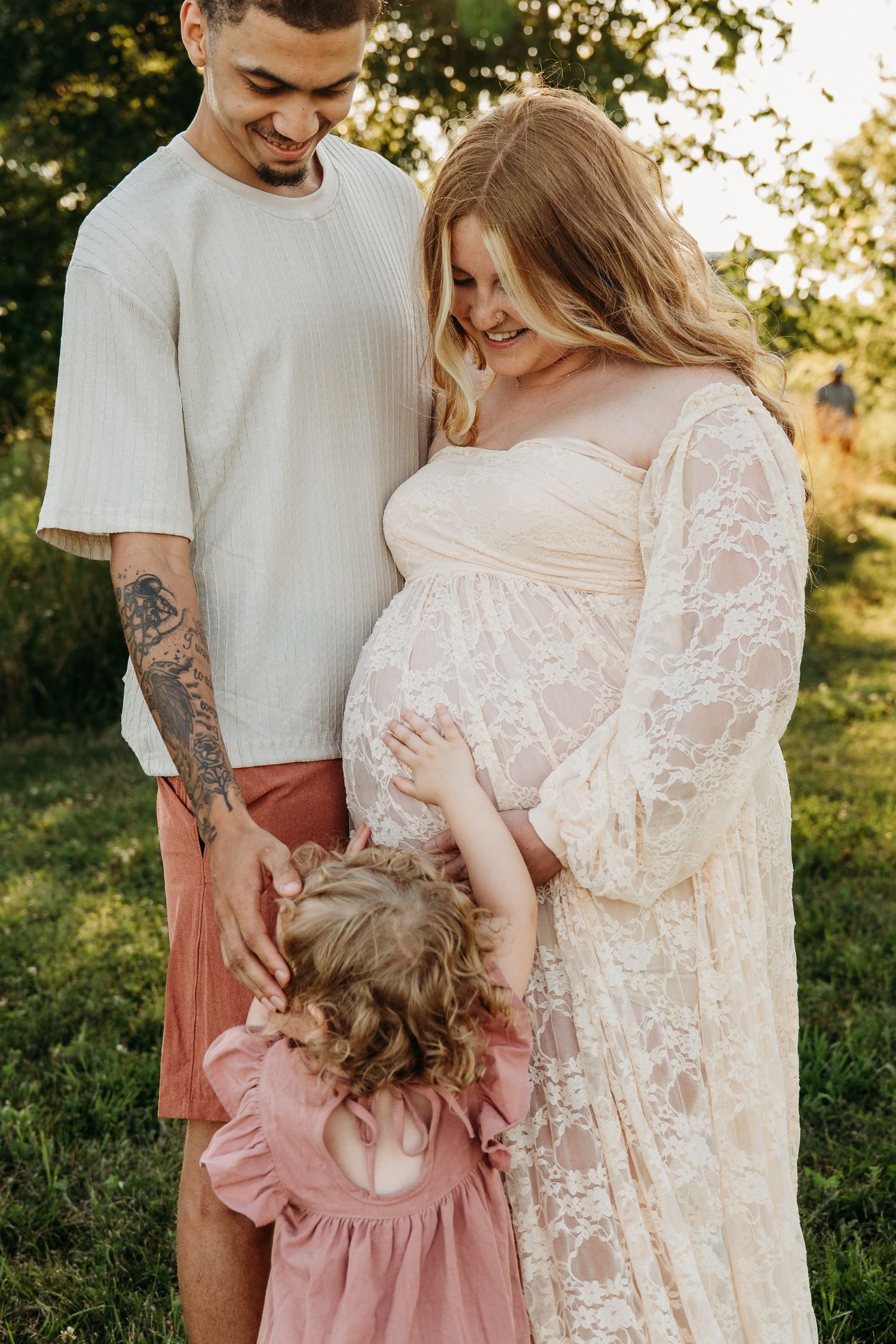 A pregnant woman in a lace dress stands with family outdoors during a sunset maternity photoshoot.