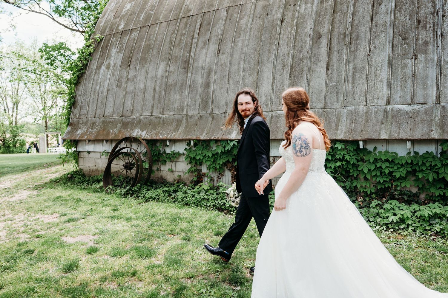 A wedding couple walking together on grass near a rustic wooden barn on a spring day.