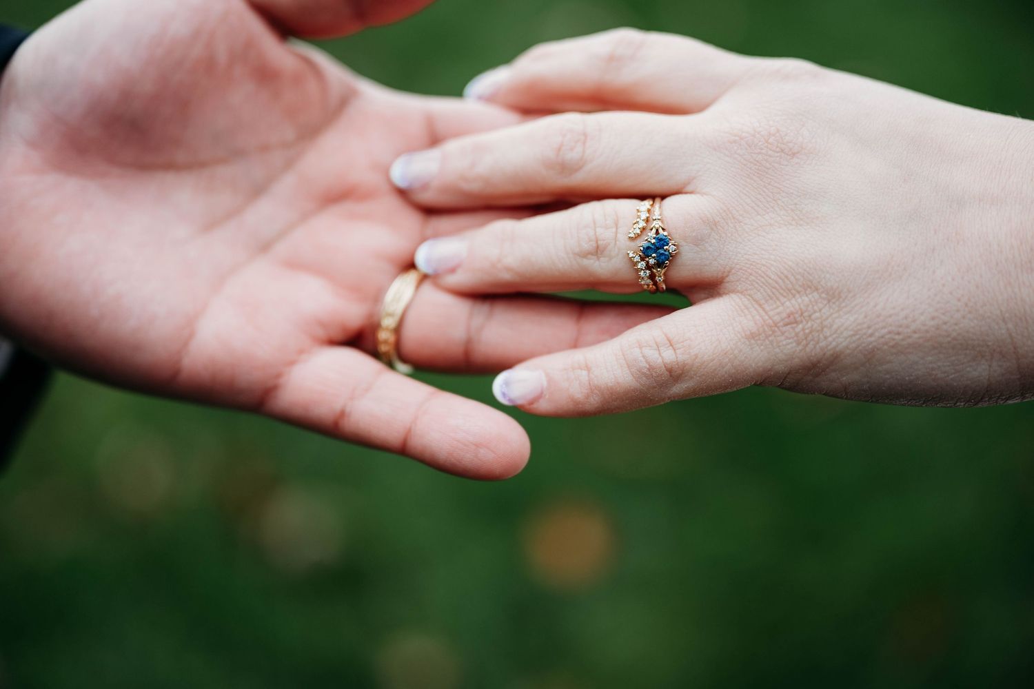 Close up shot of two hands wearing wedding rings against a blurred green natural background.