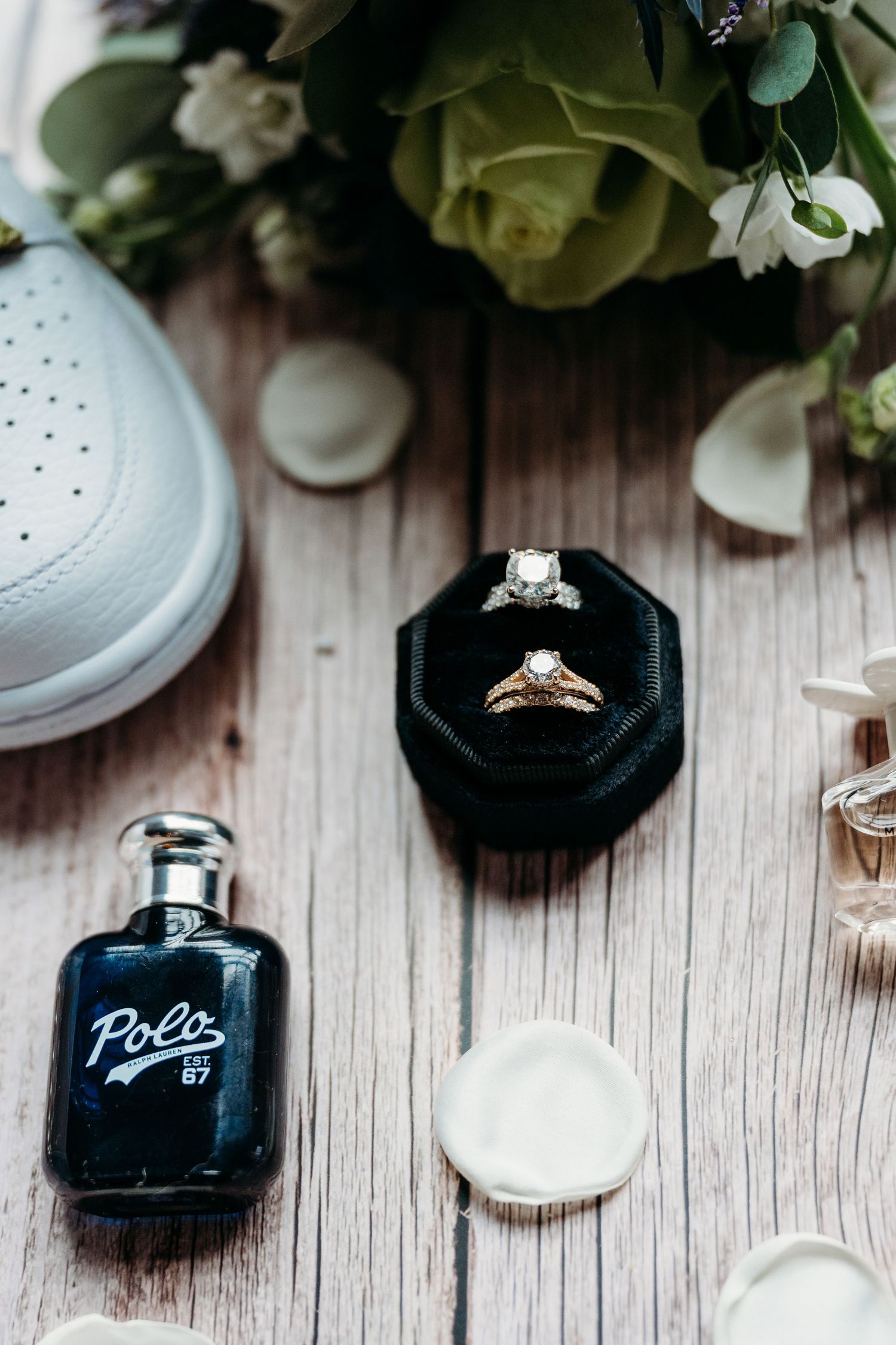 Wedding rings and perfume bottles arranged on rustic wooden surface with white flowers.