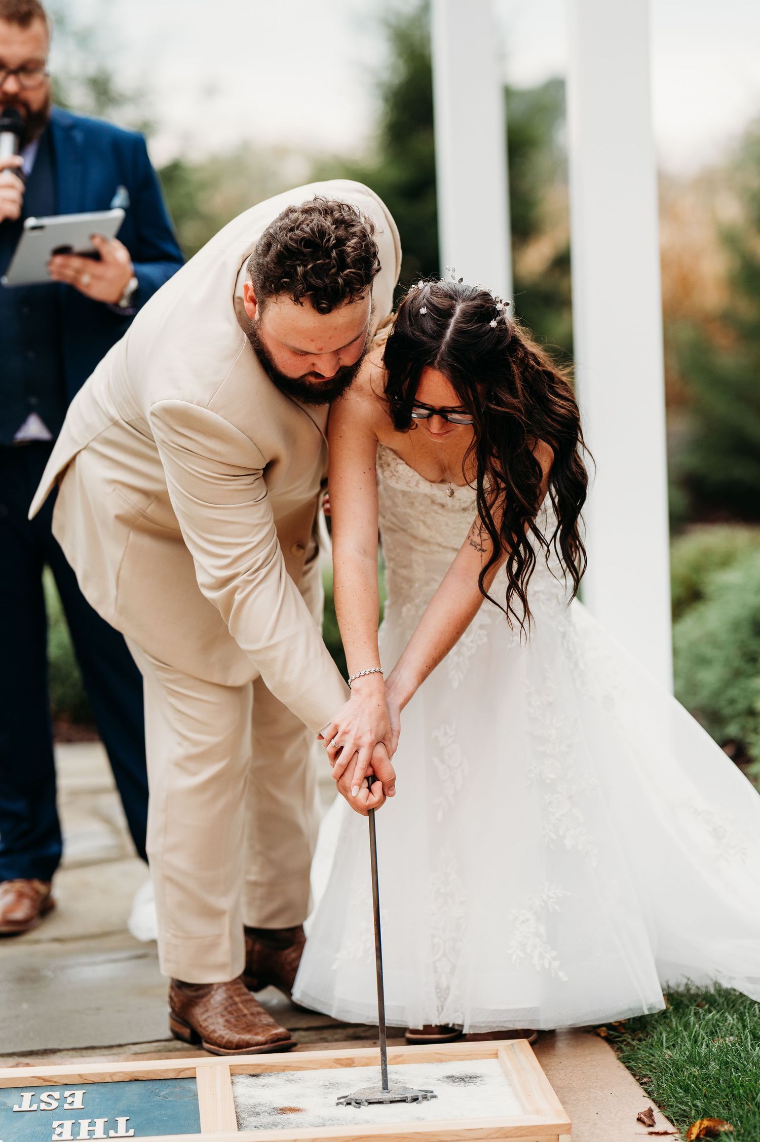 Wedding couple participates in a ceremonial signing ritual on a white surface outdoors.