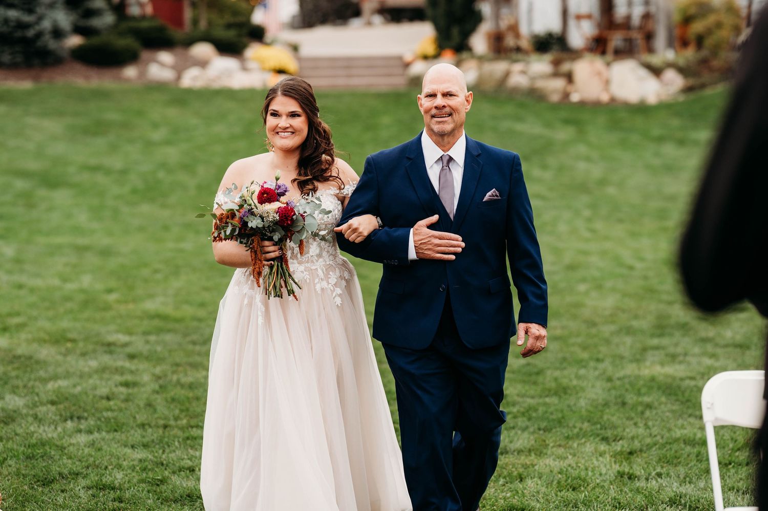 A bride in a flowing white gown walks arm in arm with an older gentleman in a navy suit during an outdoor ceremony.