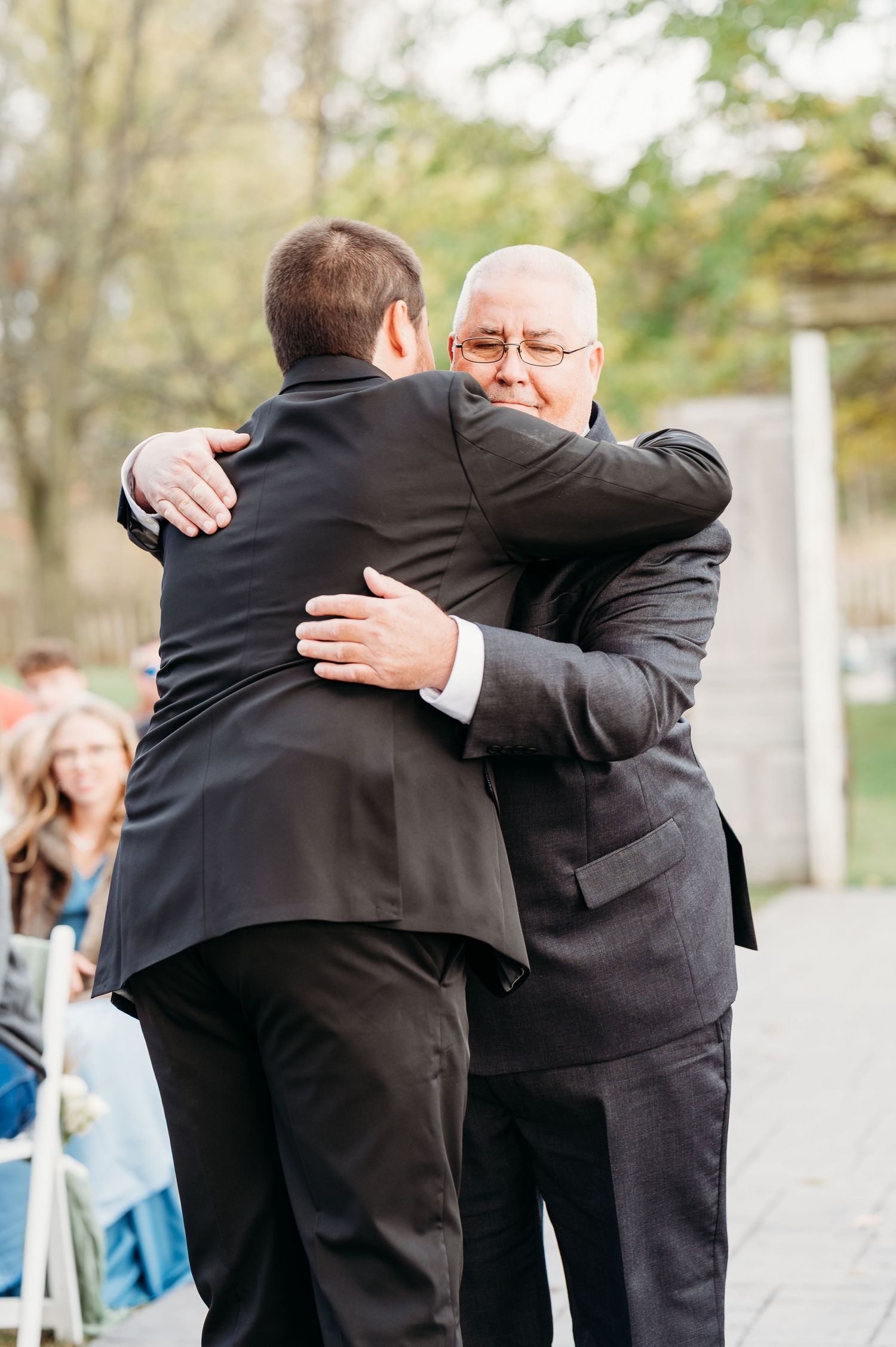 Two men in dark suits share an emotional embrace at an outdoor event.