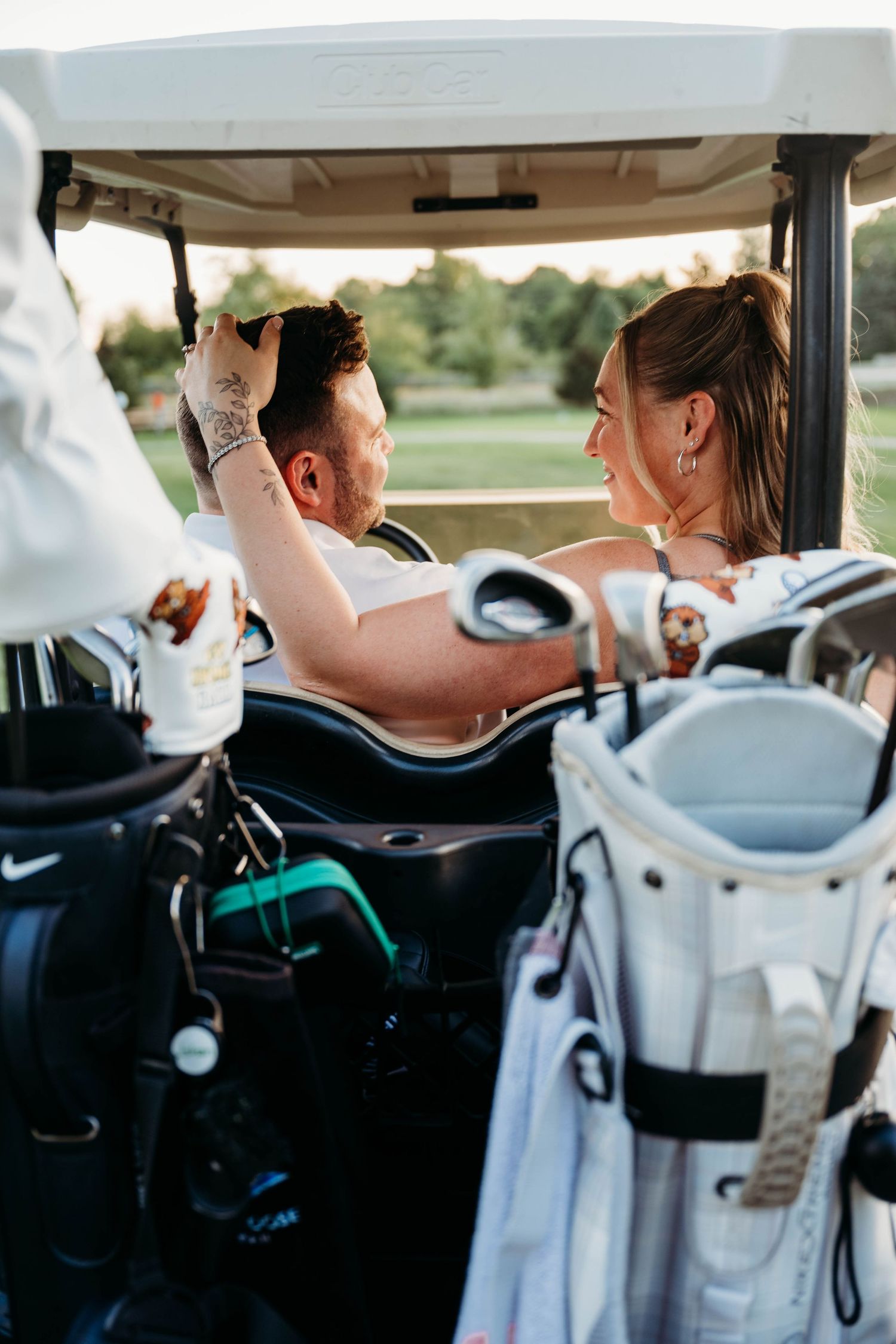 A couple shares an intimate moment while riding in a golf cart on a sunny day at a golf course.