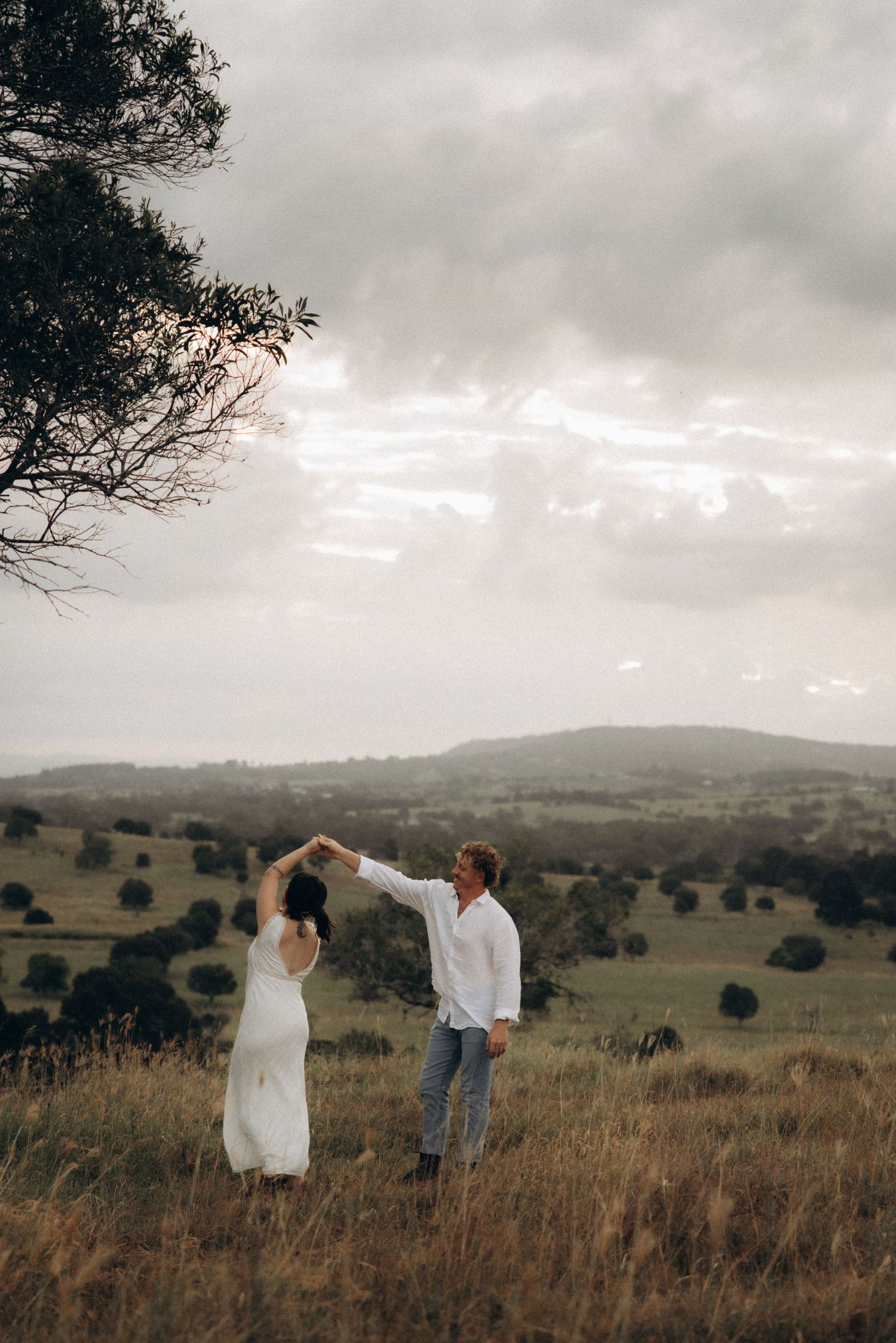 A couple dances together in a scenic field with rolling hills and moody clouds in the background.