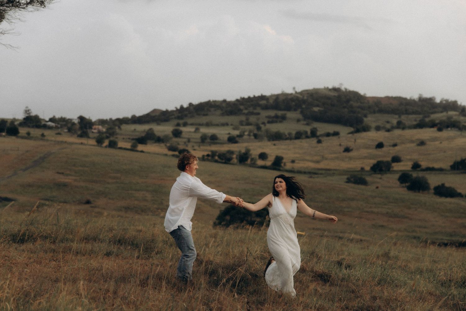 Couple running through a scenic grassy field with rolling hills in the background.