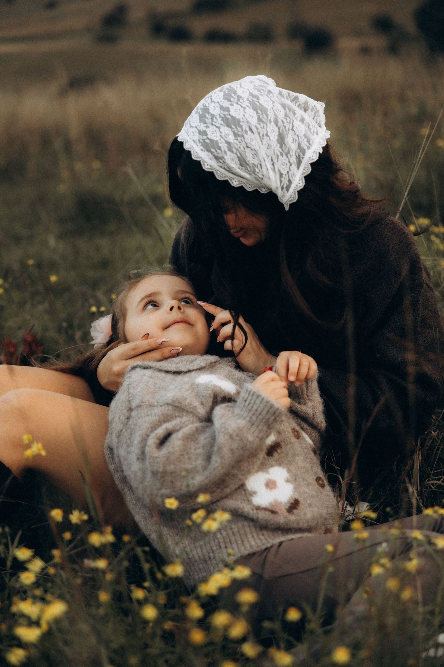 A mother wearing a white lace headscarf sits in a flower field, tenderly interacting with her young daughter.