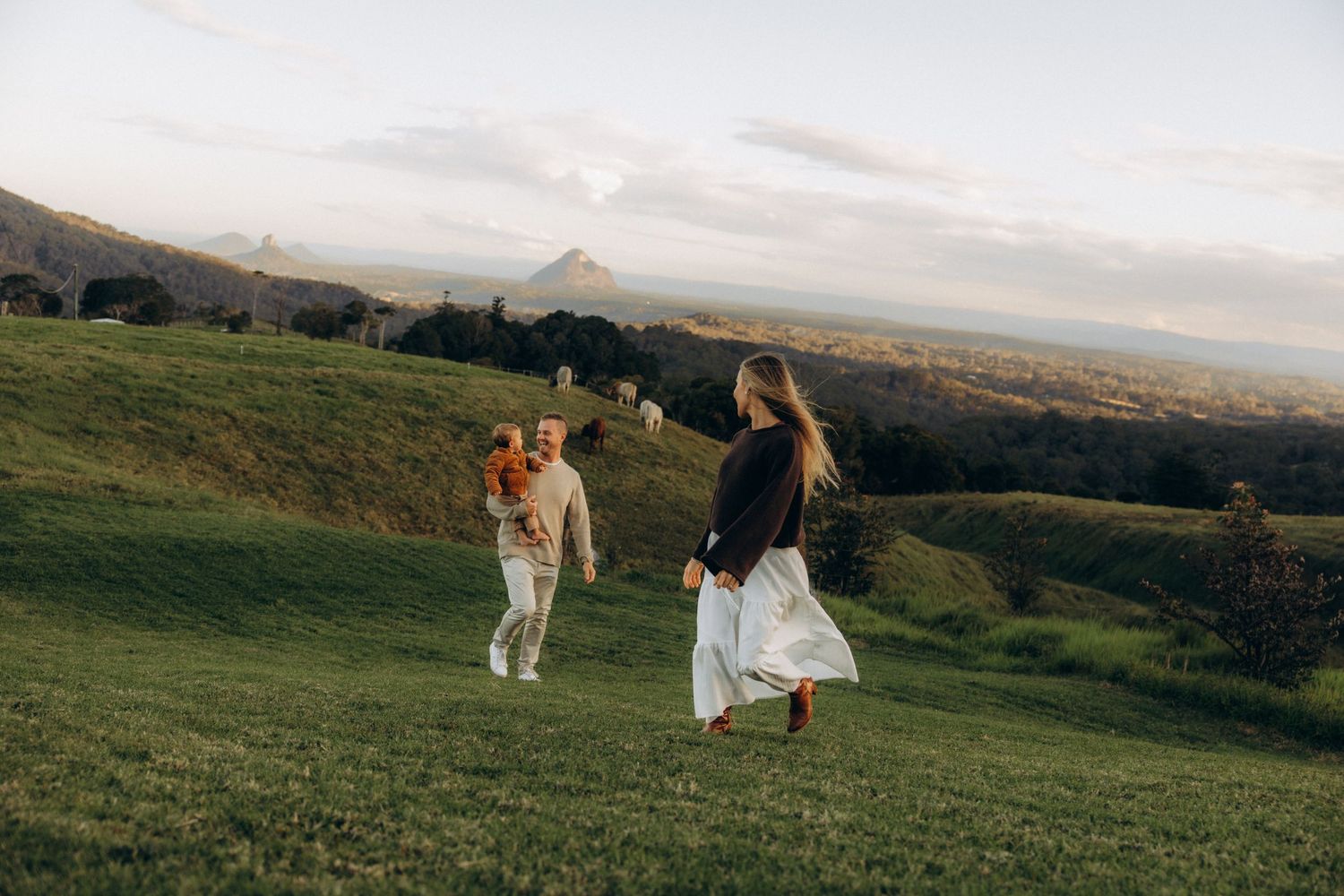 Couple walking on lush green hillside at golden hour with mountain views in background.