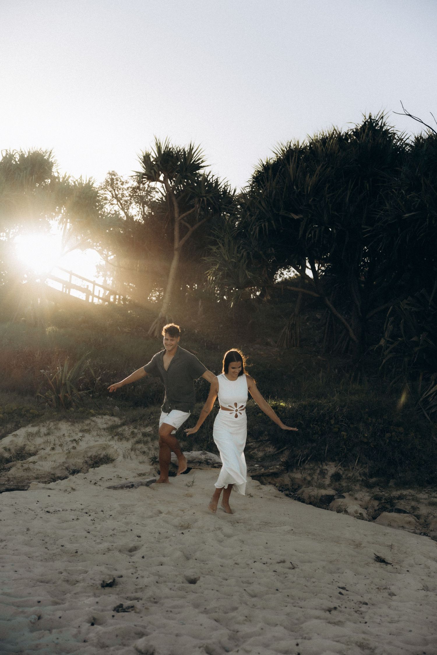 Couple holding hands walking on sandy beach path at golden sunset with tropical trees in background.