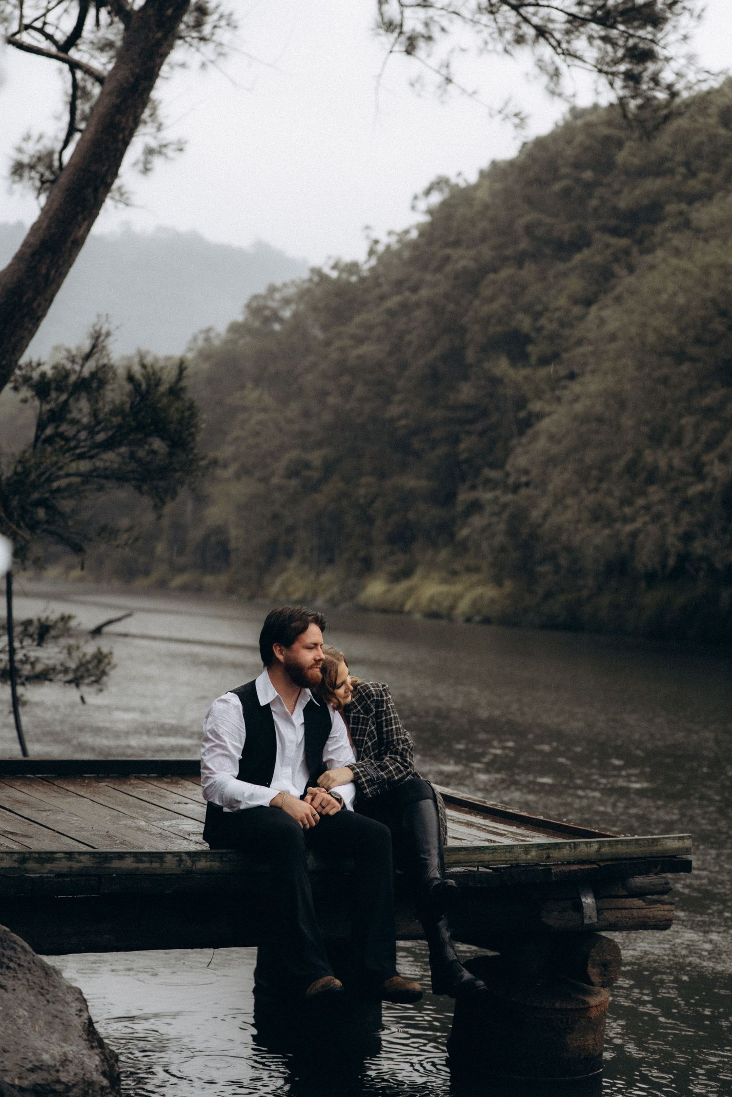 Couple sitting on wooden dock by misty lake surrounded by mountains and trees.