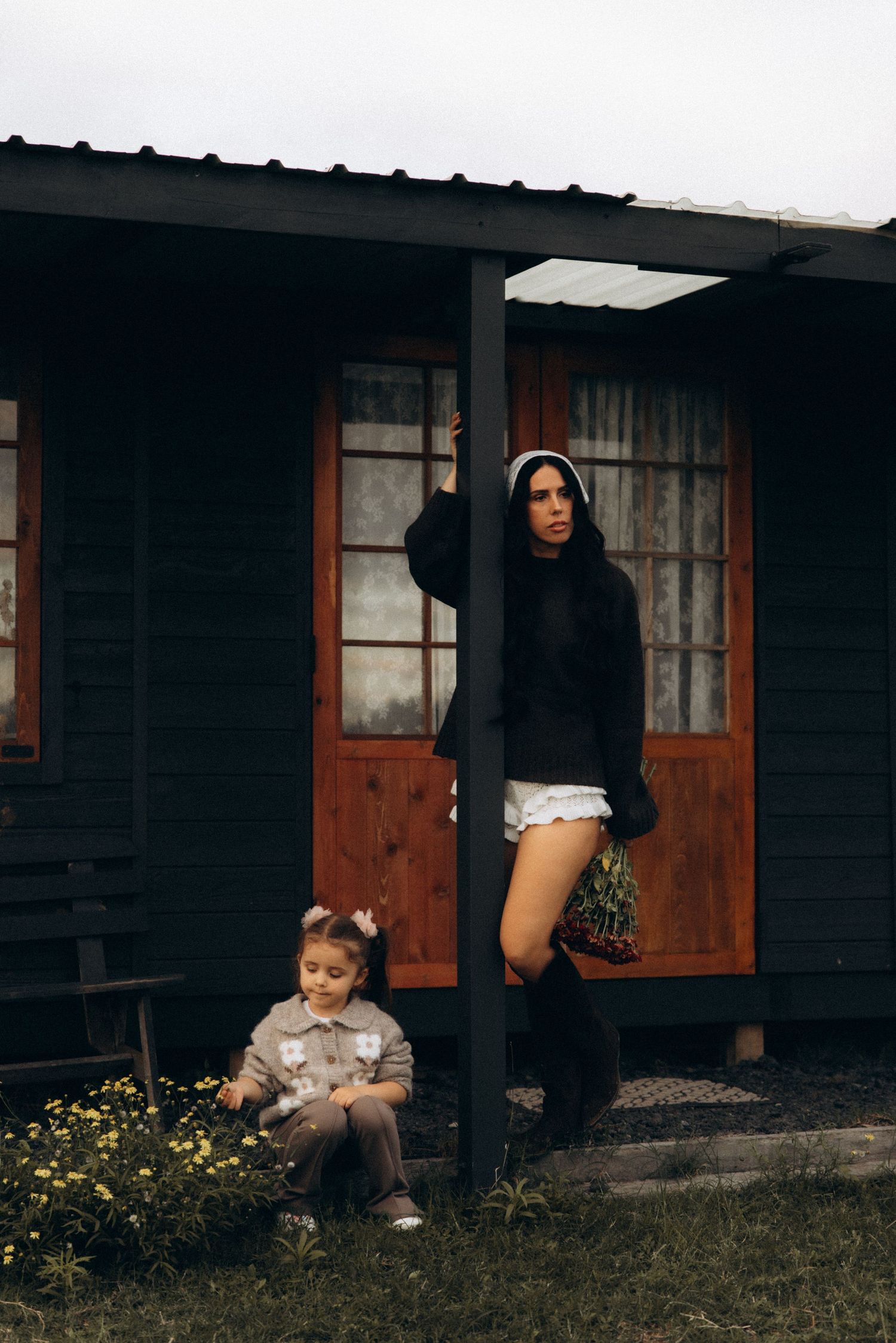 Woman in black hoodie and shorts leaning on porch post beside young girl sitting near flowers outside dark wooden house.