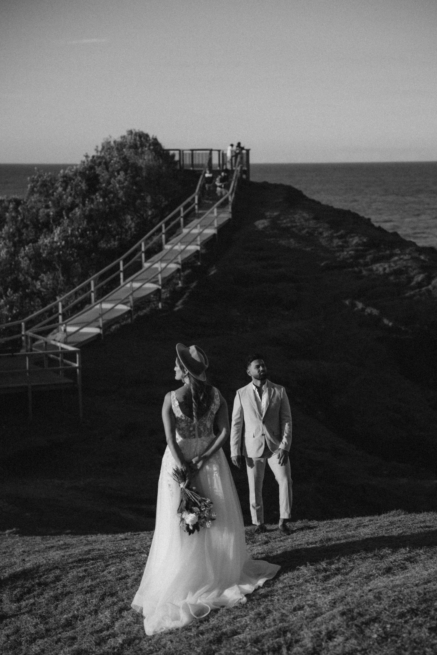 A couple in wedding attire walks along a coastal path with ocean views at sunset.