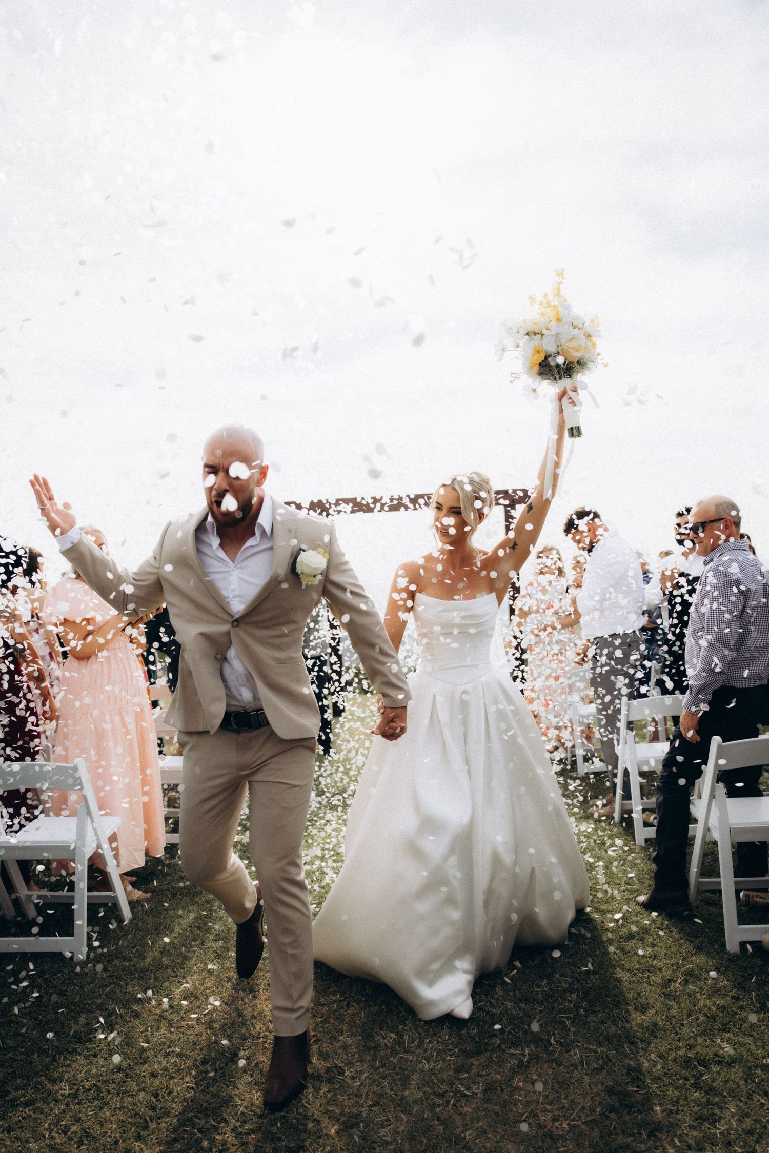 Wedding party celebrating with confetti toss outdoors against cloudy sky.