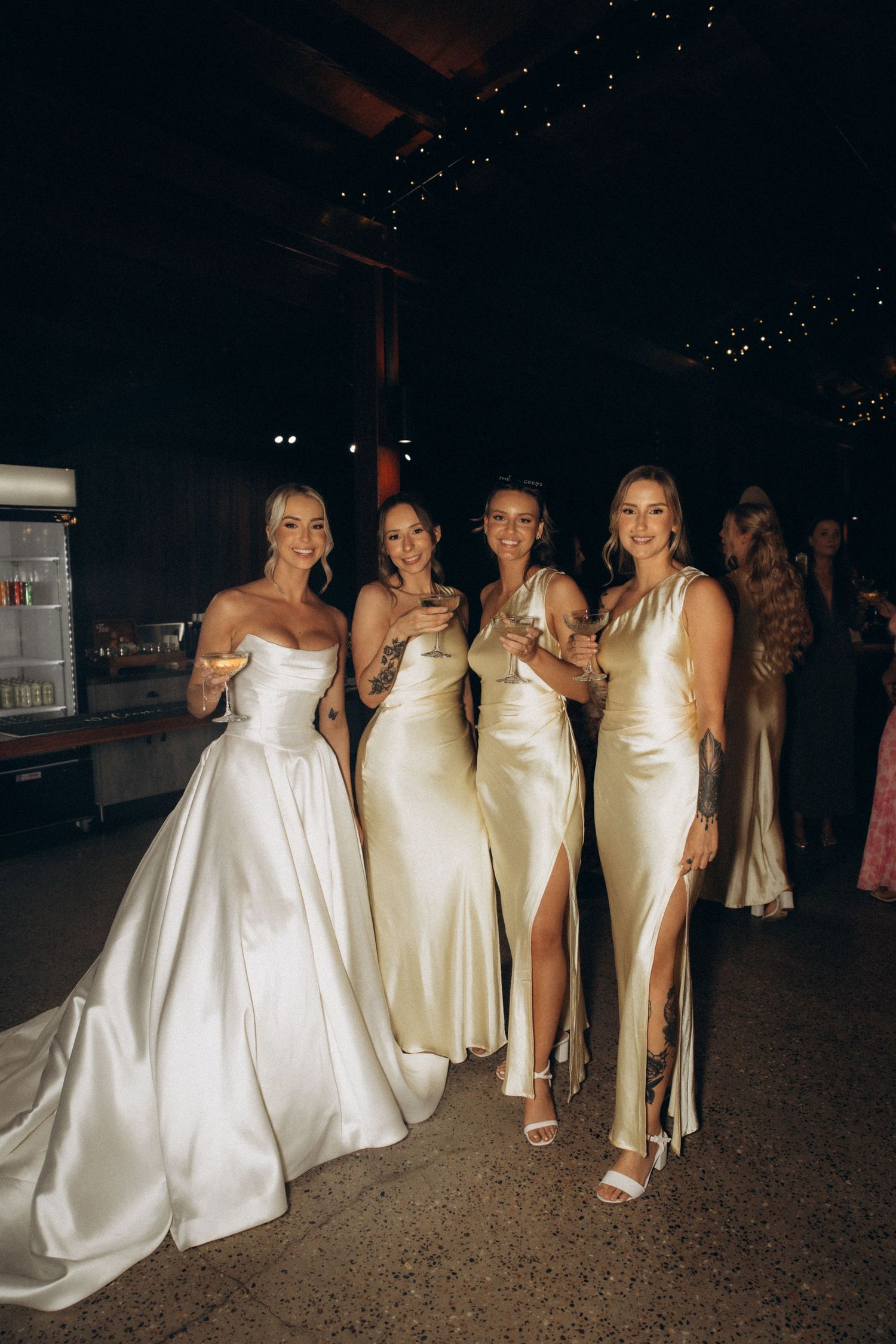 Wedding party members in elegant white and cream dresses pose together at an evening celebration.