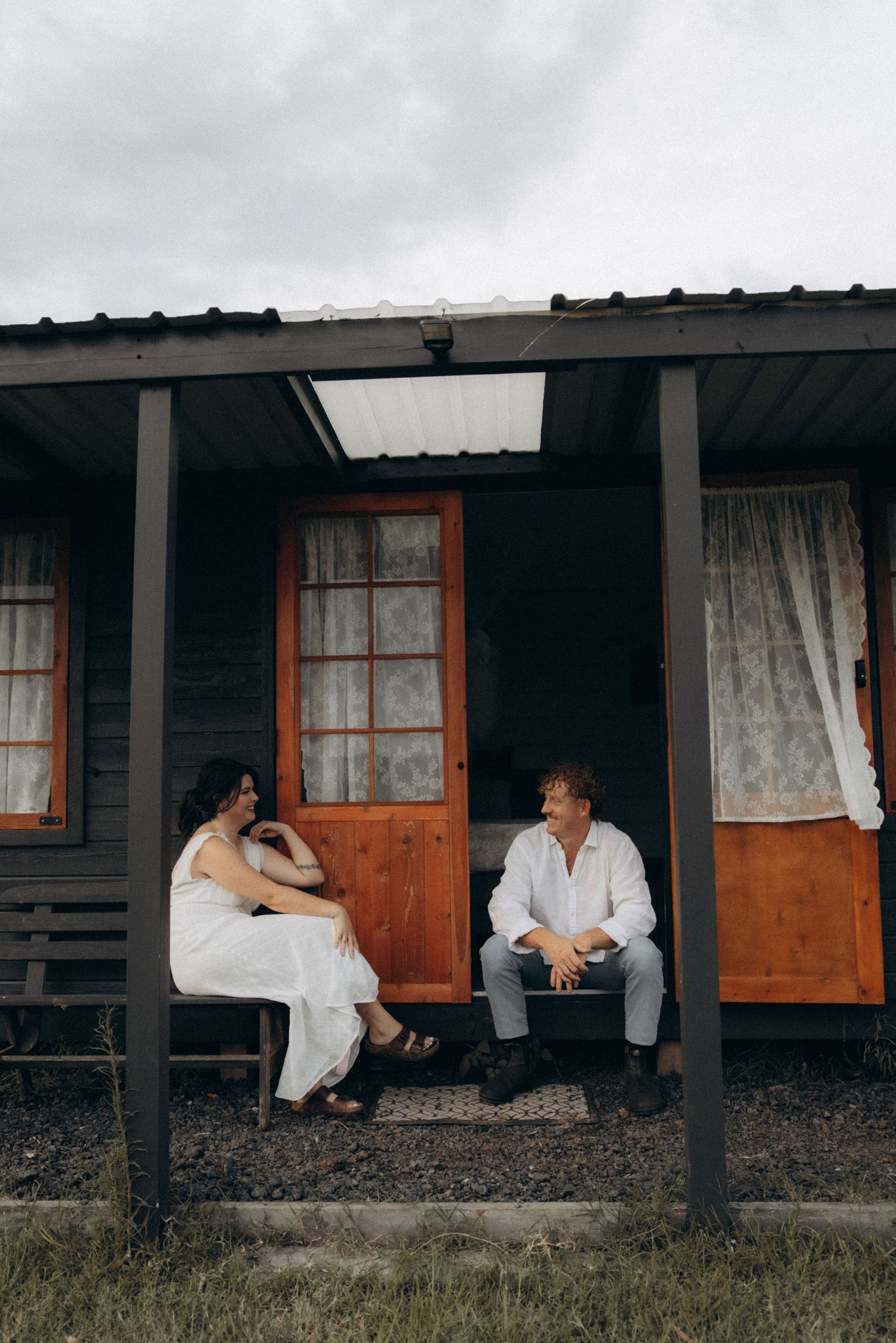 A couple sits together on the porch of a rustic cabin with wooden walls and lace curtains.