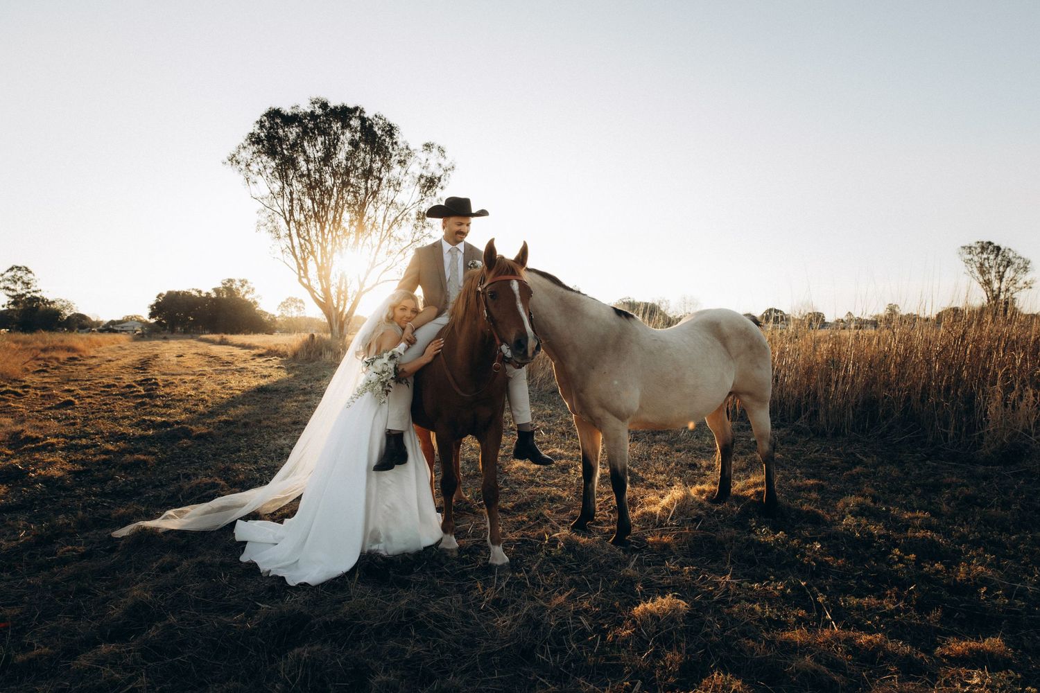 Bride and groom standing with two horses in a golden field at sunset during their rustic country wedding.