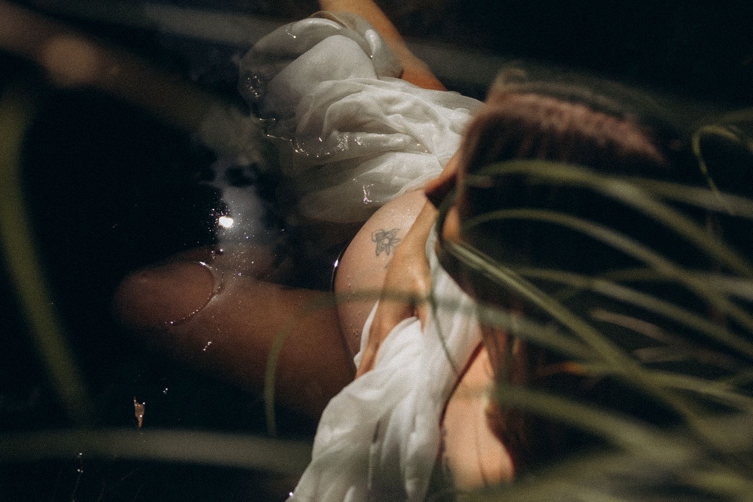 Person in white fabric lying near water and tropical plants in a moody, artistic outdoor setting.