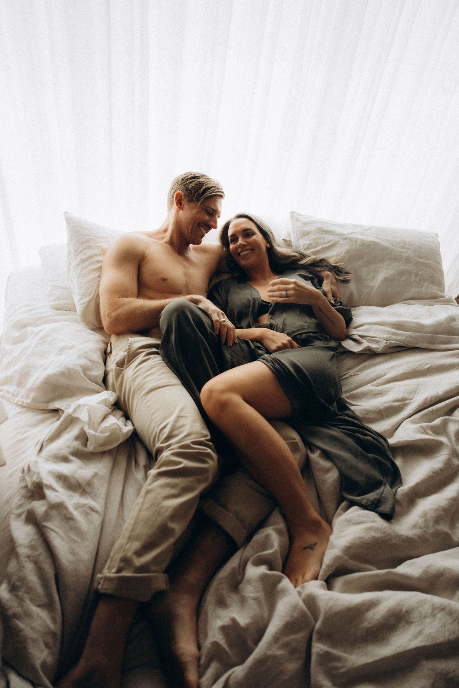 A couple relaxes together on white bedding in natural window light.