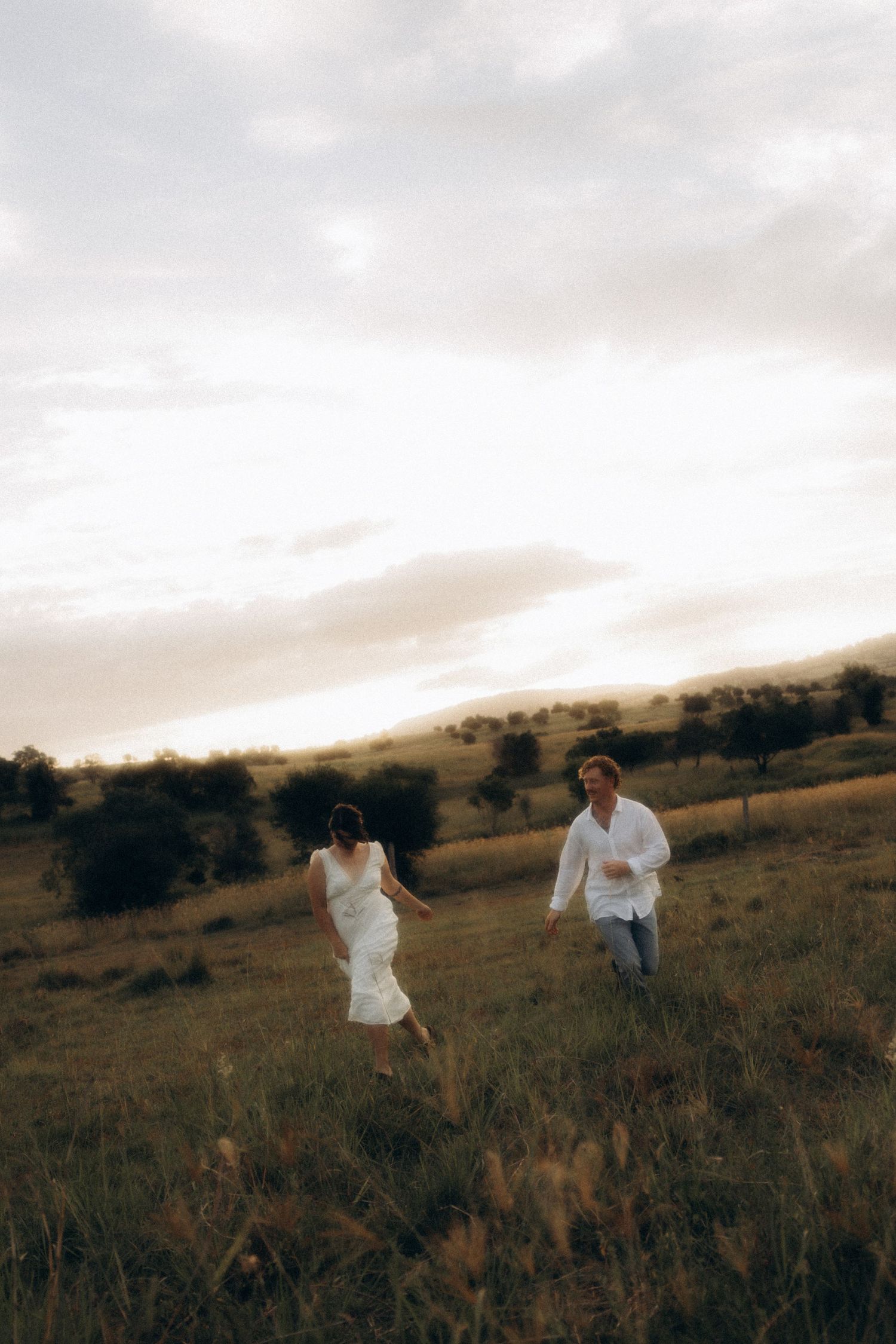 Couple in white clothing walking through a golden field at dusk.